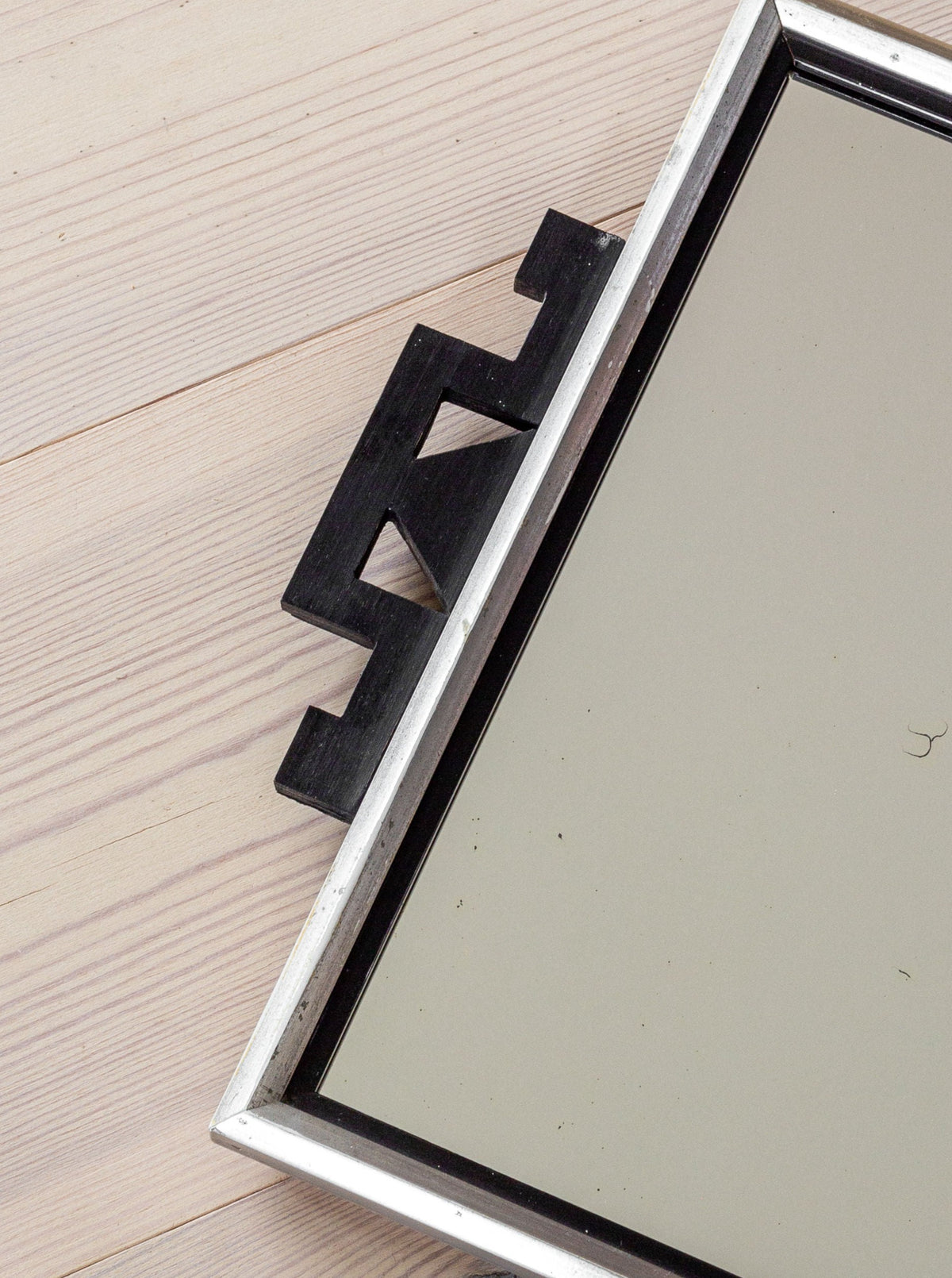 A vintage Spigel mirror from the 1930s, known as the Swedish Art Deco Mirror, with a silver frame and a distinctive black geometric handle, rests on a light wooden surface. The handle features a unique zigzag design inspired by Art Deco, adding an elegant touch.