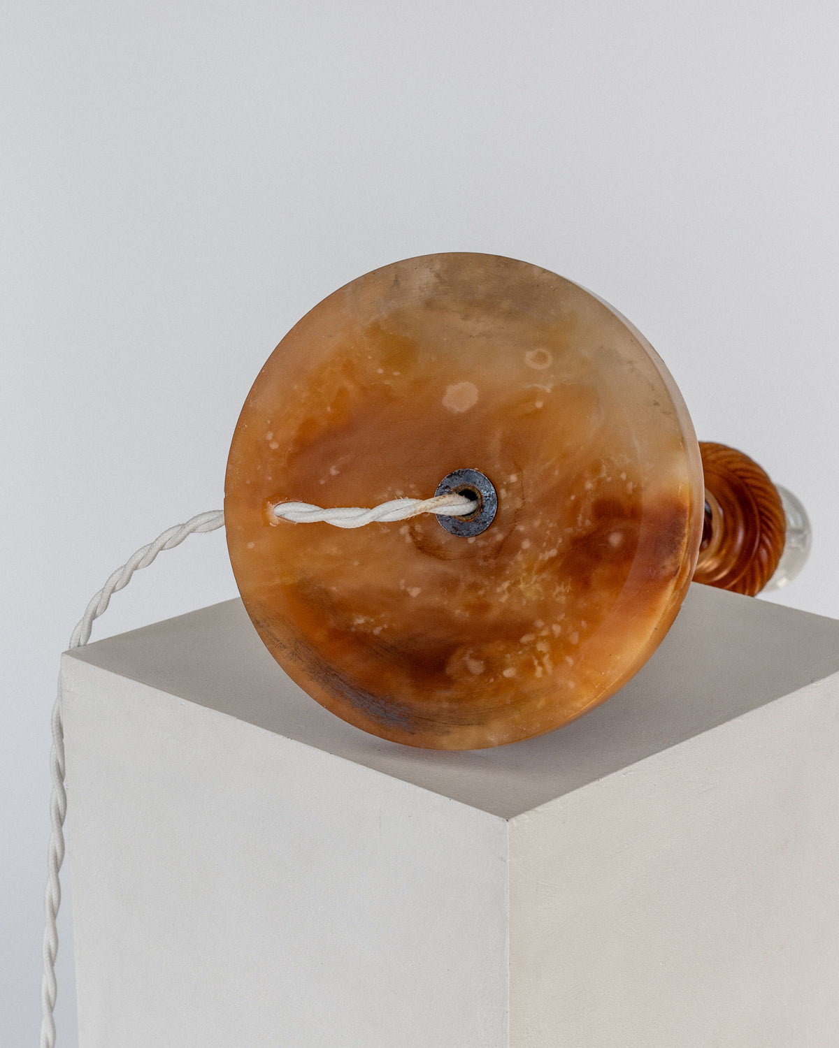 The Antique Alabaster Table Lamp, C. 1900 by Spigel exudes a classic and timeless aesthetic with its round, amber-colored base featuring a marbled texture. Displayed on its side atop a white pedestal, the hand-carved alabaster base radiates elegance. A cord extends from the center of the base, draping gracefully in a loop.