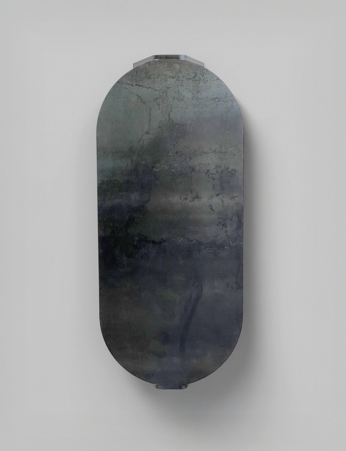 The 'Tani' Coffee Table by Modan features a vertically oriented, oval-shaped steel top with a dark, reflective surface accented by abstract, cloudy gray, blue, and green patterns, set against a plain light gray background.