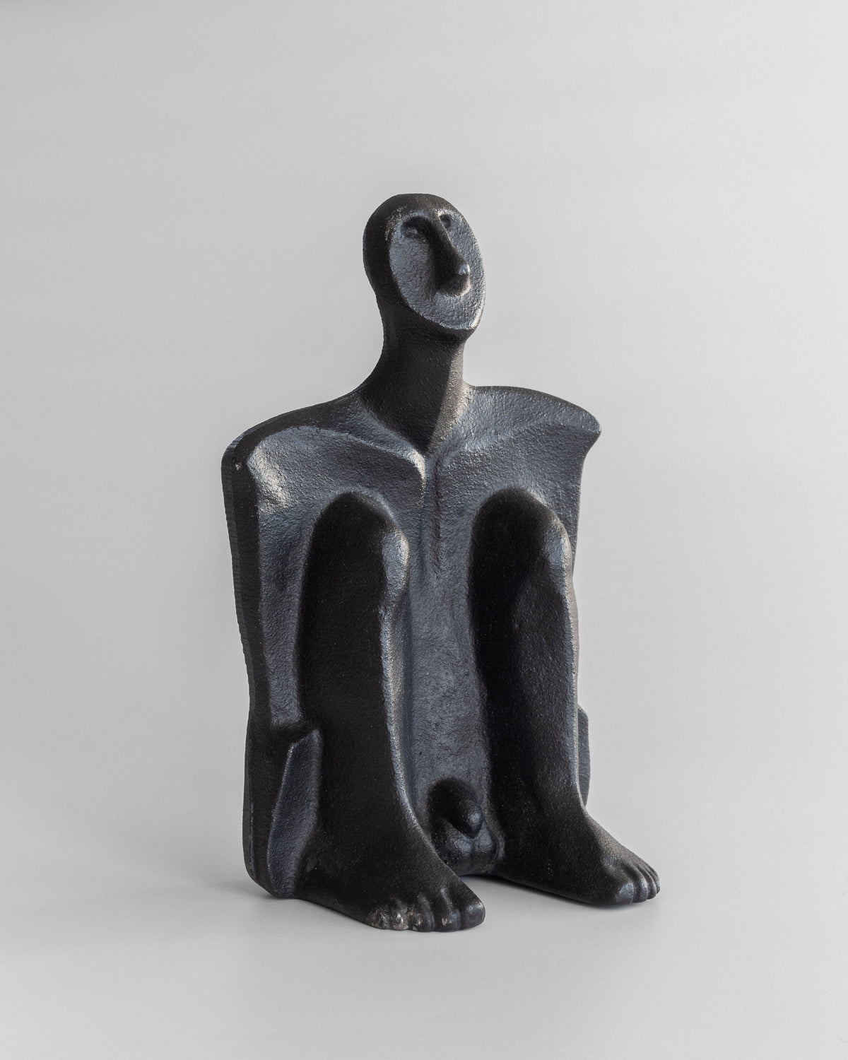 A black abstract seated figure by Spigel, crafted in cast iron by Marita Norin, rests on a light gray background. Its stylized features and exaggerated limbs make it ideal for artistic bookends.