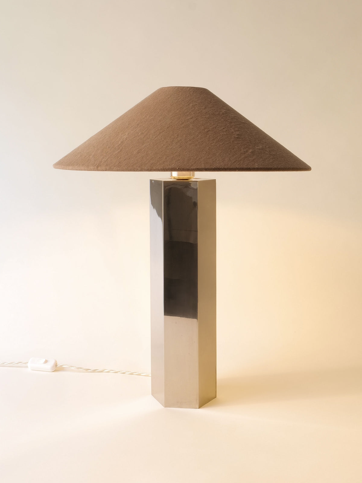 The Grand Hexagonal Lamp by Collection apart features a tall stainless steel base and a wide brown conical fabric shade, bringing stylish 1970s retro flair to any space. Displayed on a light surface against a neutral background.