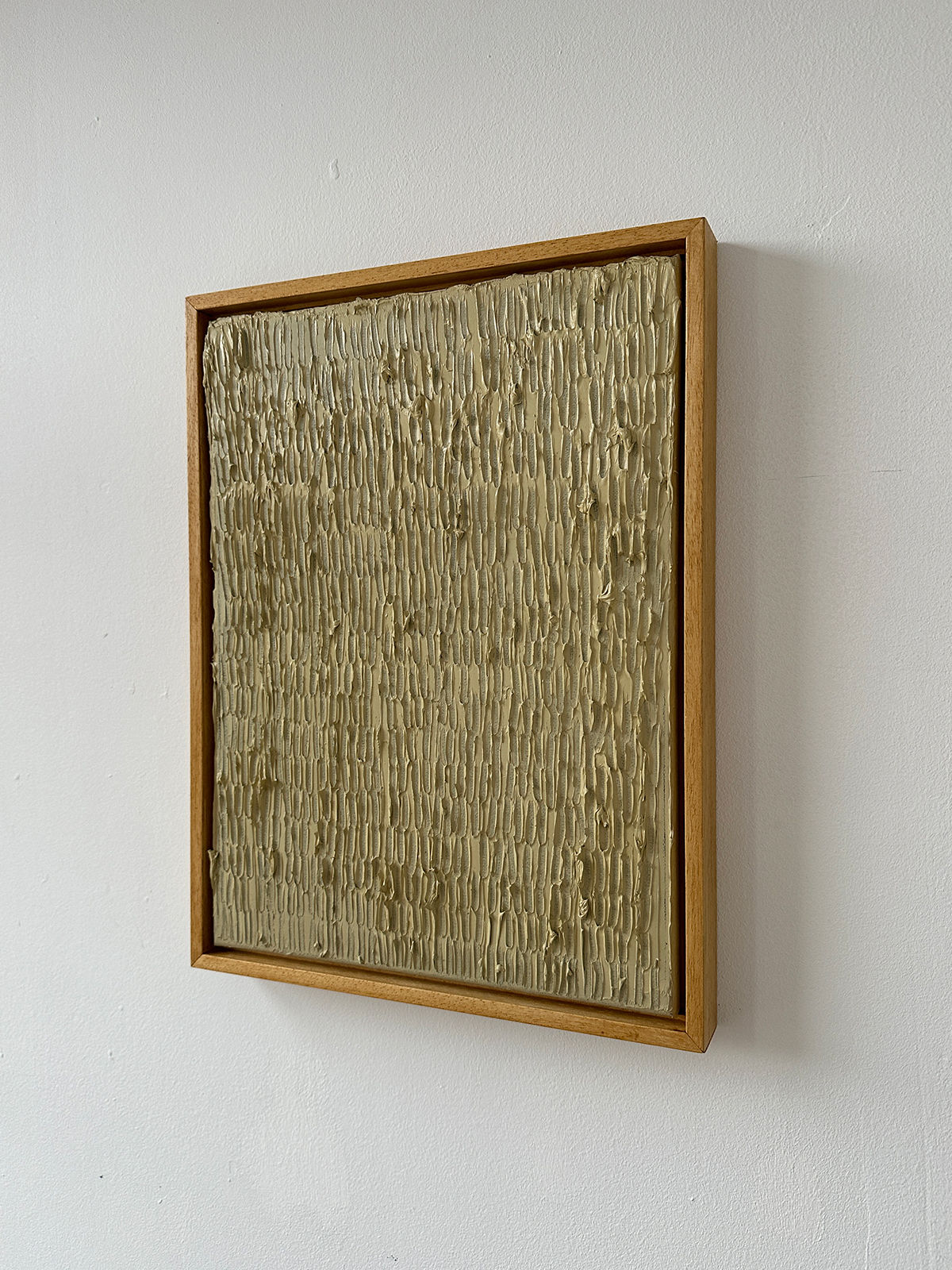 The "Buff Titanium" painting by Roi Elmaliah is a textured, square artwork showcasing vertical ridges on a beige surface. Crafted with oil on canvas, it is surrounded by a thin wooden border and mounted on a white wall.