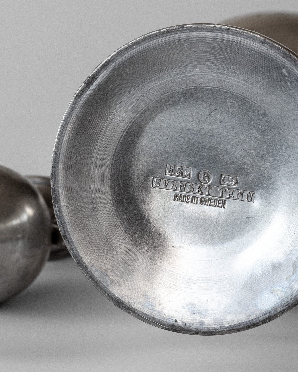 Close-up of a metal object with stamped text reading "ESM StG LD Svenskt Tenn Made in Sweden." Crafted by Ernst Svedbom, this piece is part of Spigel's 1953 collection, including neoclassical metal vases, showcasing a shiny metallic surface reminiscent of elegant pewter designs.