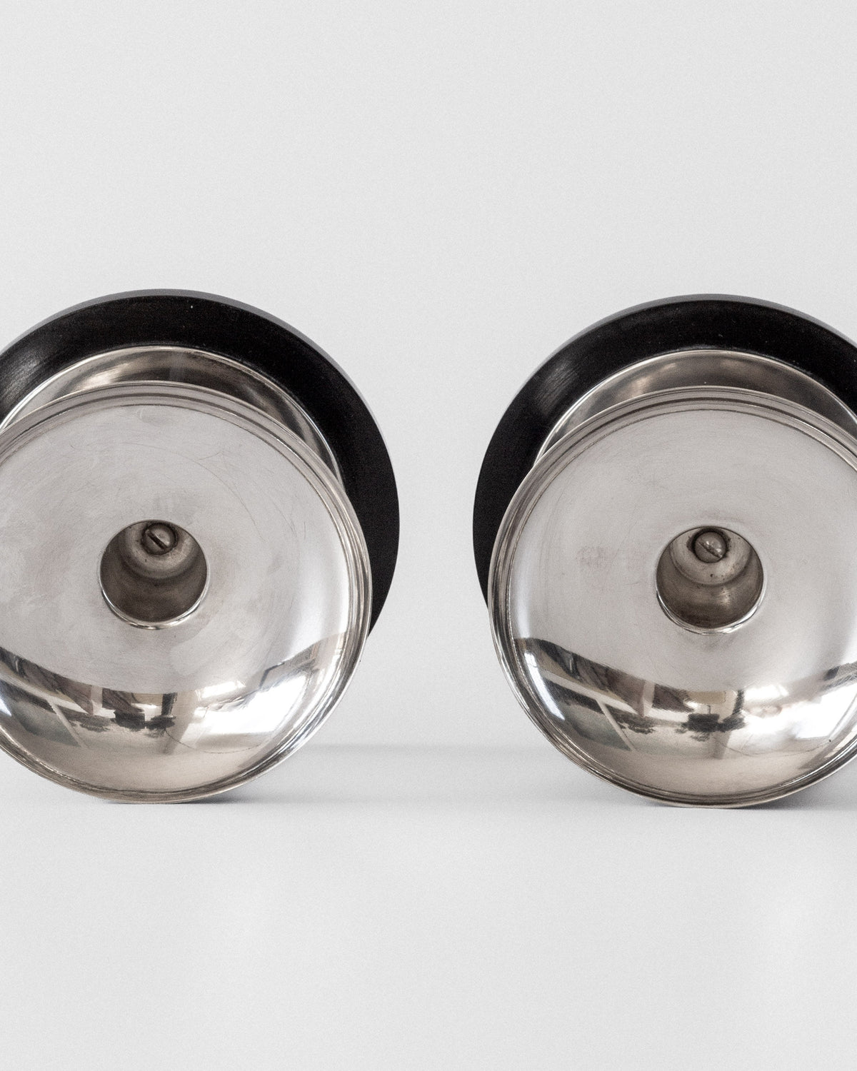 Two Modernist Sterling Silver Candleholders by Spigel (1934) with black rims are set side by side on a white surface, viewed from above. Their symmetrical, circular shapes and central protrusions showcase modernist design.