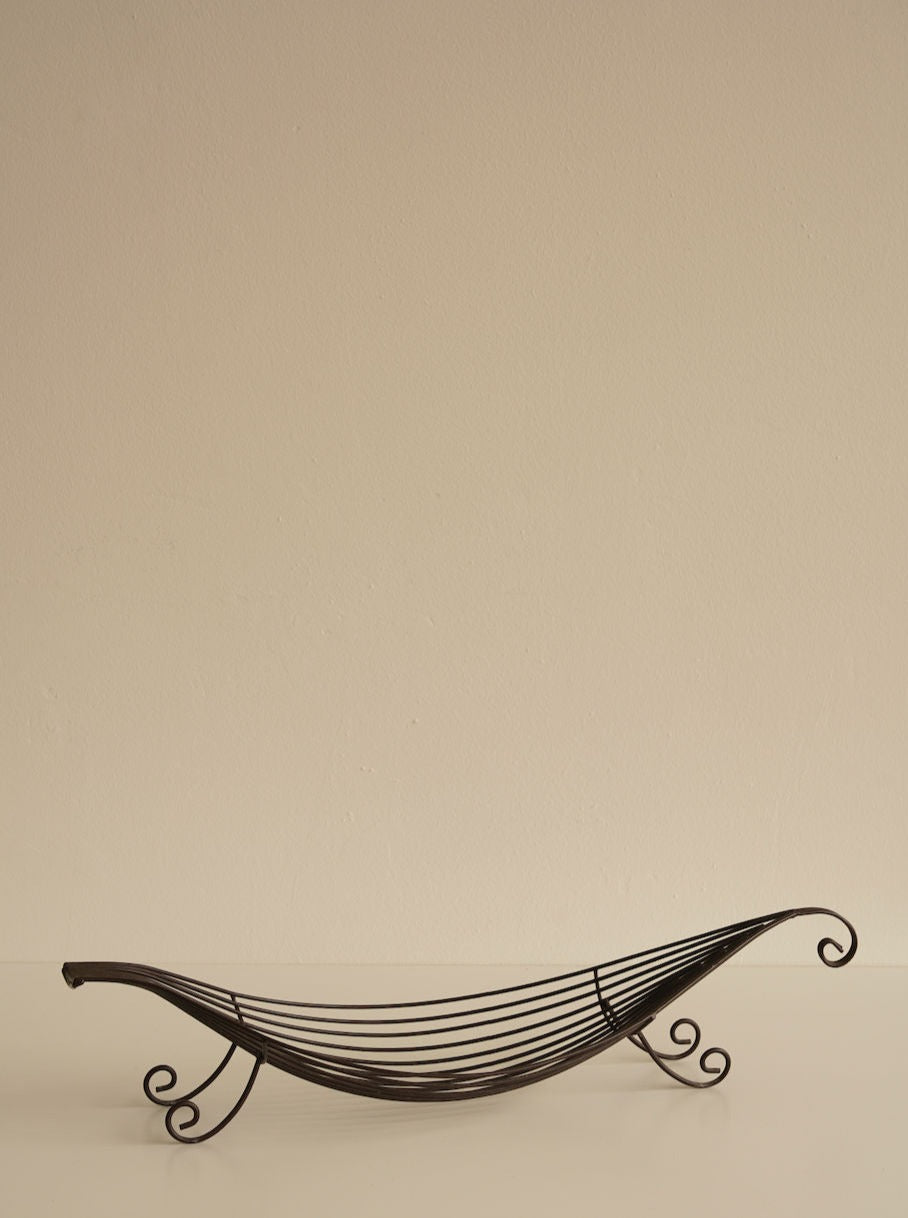 An Iron Swirl Fruit Bowl by Les Objets Amsterdam with an elegant, curved design sits empty against a plain beige background. The elegant fruit bowl features intricate, flowing loops and swirl accents on the ends and sides, creating a sleek and artistic look.