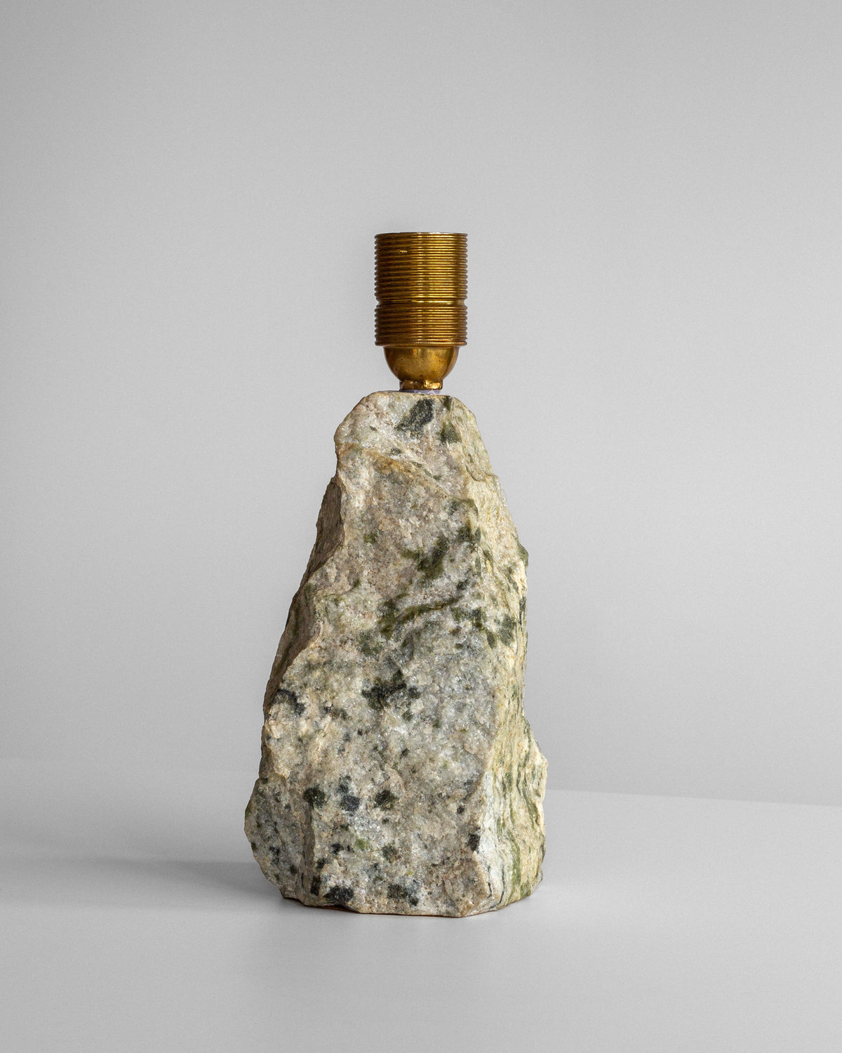 A gold-toned light socket tops the obelisk-shaped, textured rock of the Spigel Marble Stone Table Lamp, creating a unique appearance against a plain gray background.