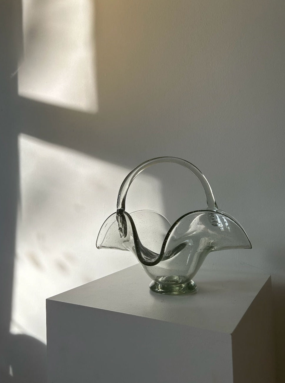 A clear glass decorative bowl with a curved, petal-like design and a handle sits on a white pedestal. The thick quality glass reflects soft sunlight from a nearby window, casting shadows on the plain wall behind it. This Les Objets Amsterdam 70's Vintage Glass Basket exudes the charm of a 70's vintage bonbonnière.