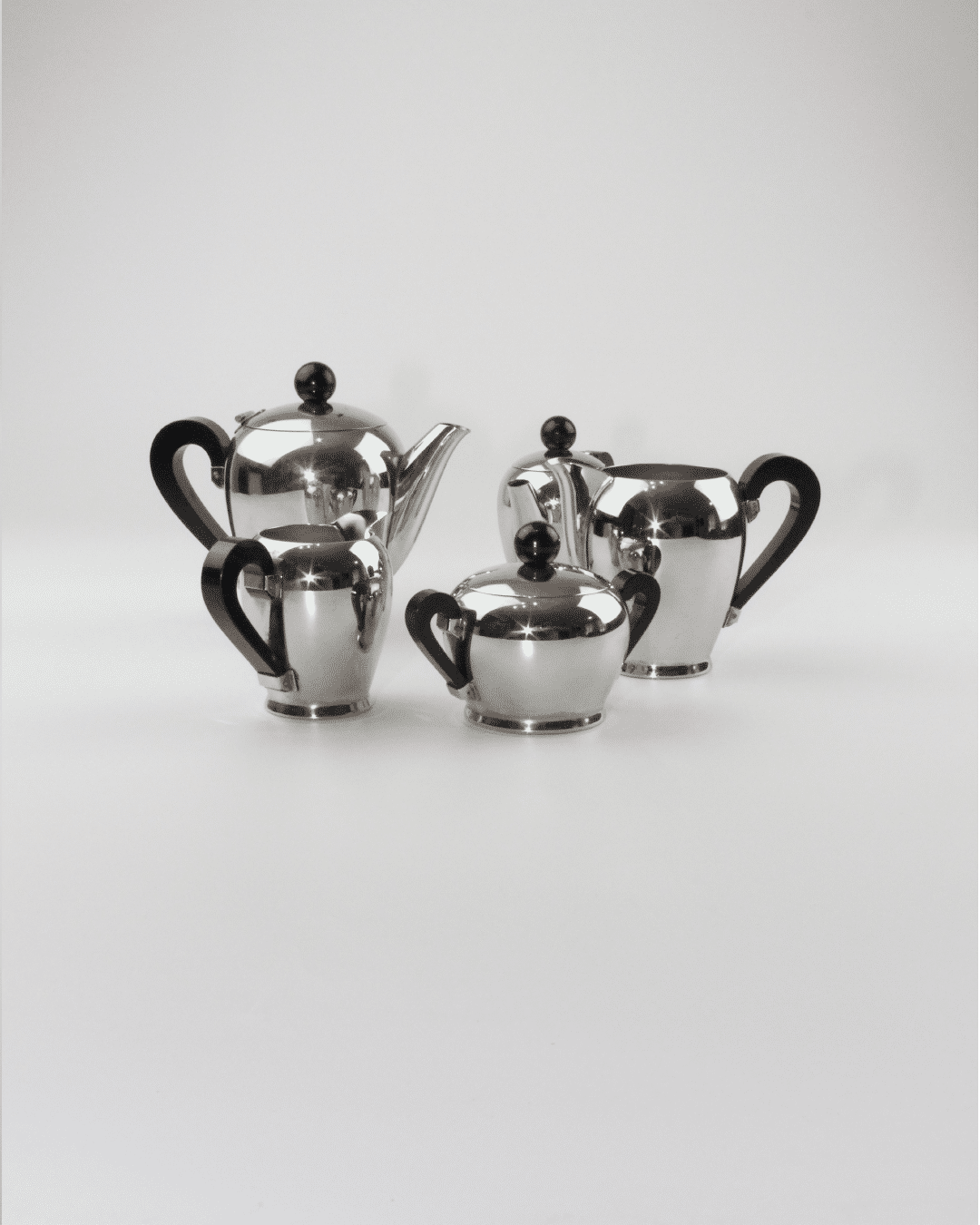 A stunning set of four polished metal teapots and pitchers of varying sizes with black handles and spherical black knobs on the lids is beautifully arranged on a white surface against a plain white background. This "Bombè" Alessi Tea/Coffee Set by Boga Avante Shop showcases mid-20th-century craftsmanship with a teapot, creamer, sugar bowl, and small pitcher.