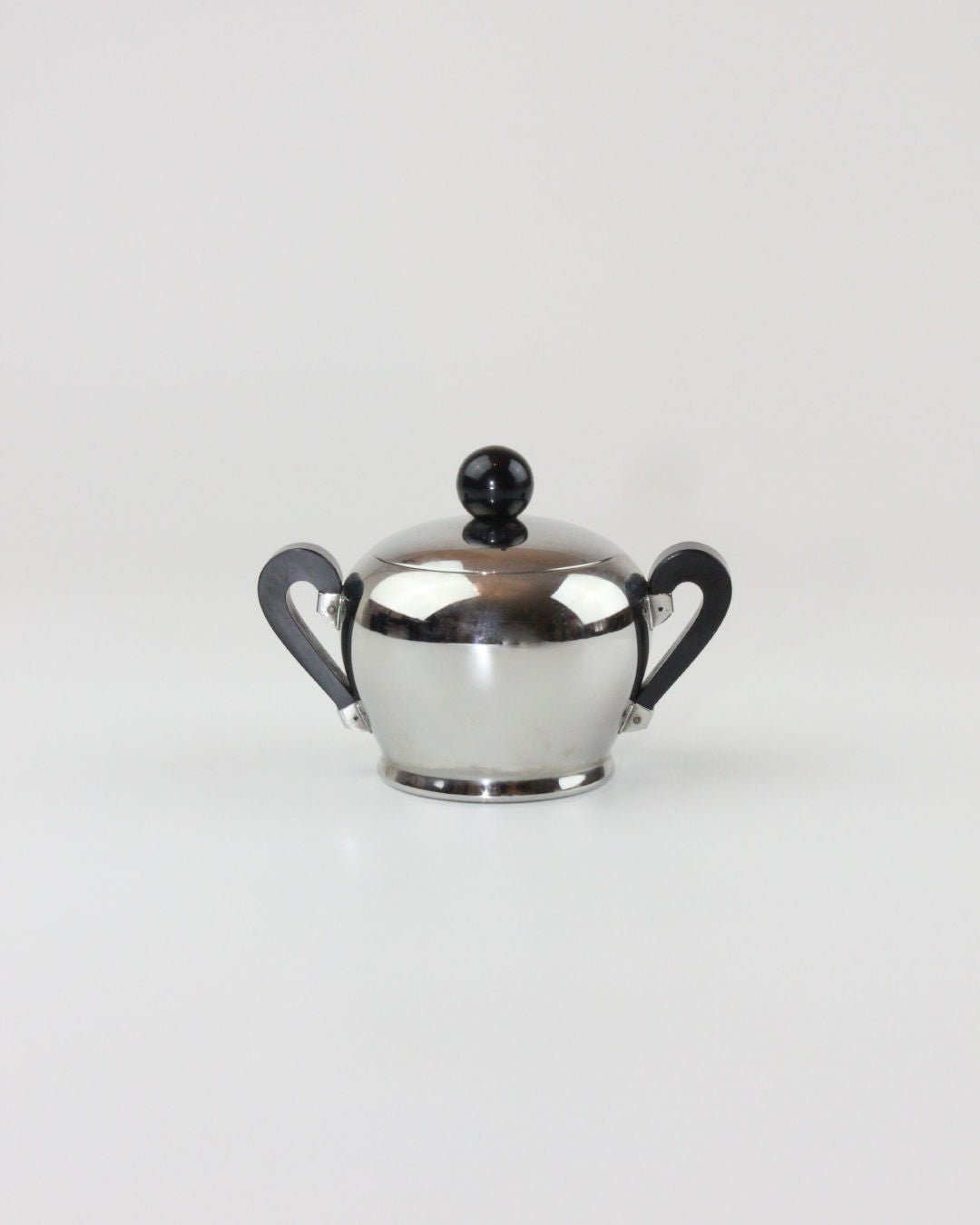 A small, shiny metal sugar bowl with a polished silver surface, two black handles, and a black knob on the lid is showcased mid-20th-century craftsmanship from the "Bombè" Alessi Tea/Coffee Set by Boga Avante Shop. The bowl is centered against a plain white background.