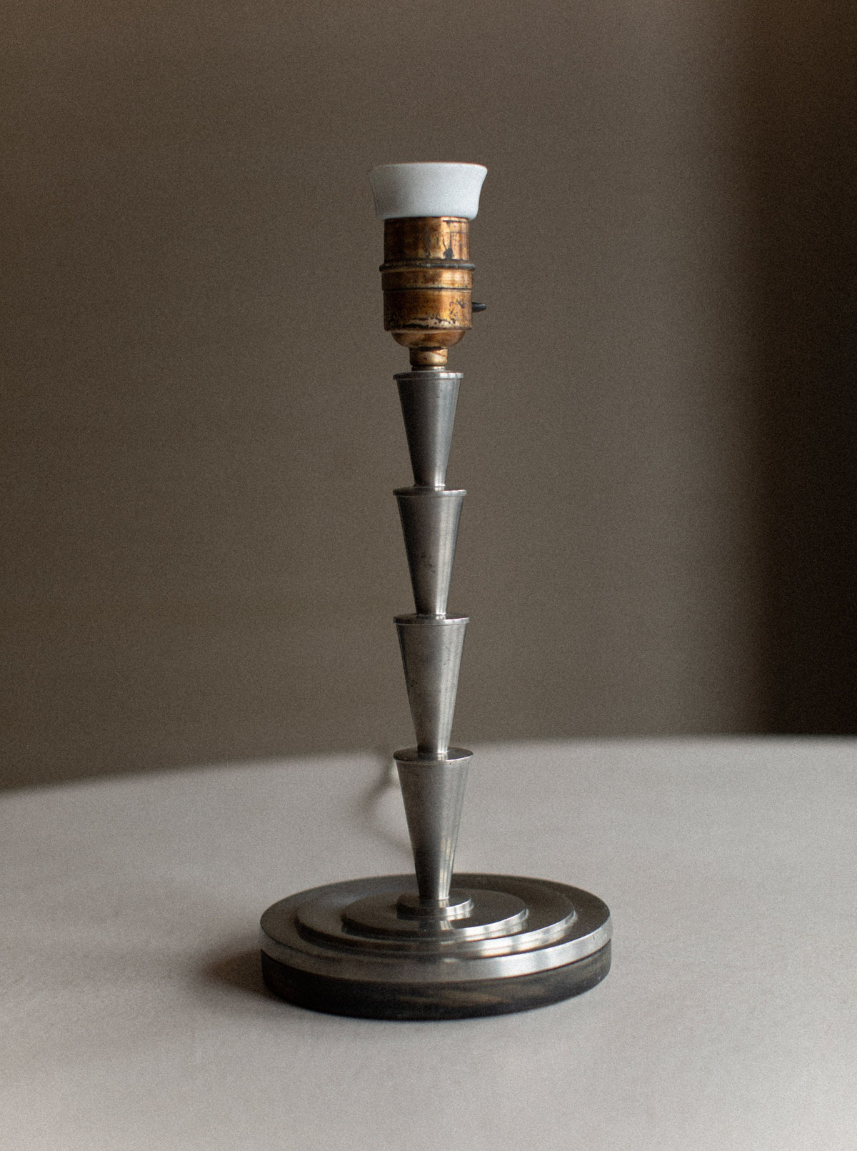 An exquisite 1920's Swedish Grace Lamp with a conical stacked design featuring three metal segments placed on a circular base. This piece of vintage decor from Out For Lunch includes a brass light fixture with a small white lamp holder. The background is blurred and neutral-colored, enhancing its timeless elegance reminiscent of an Elis Bergh design.