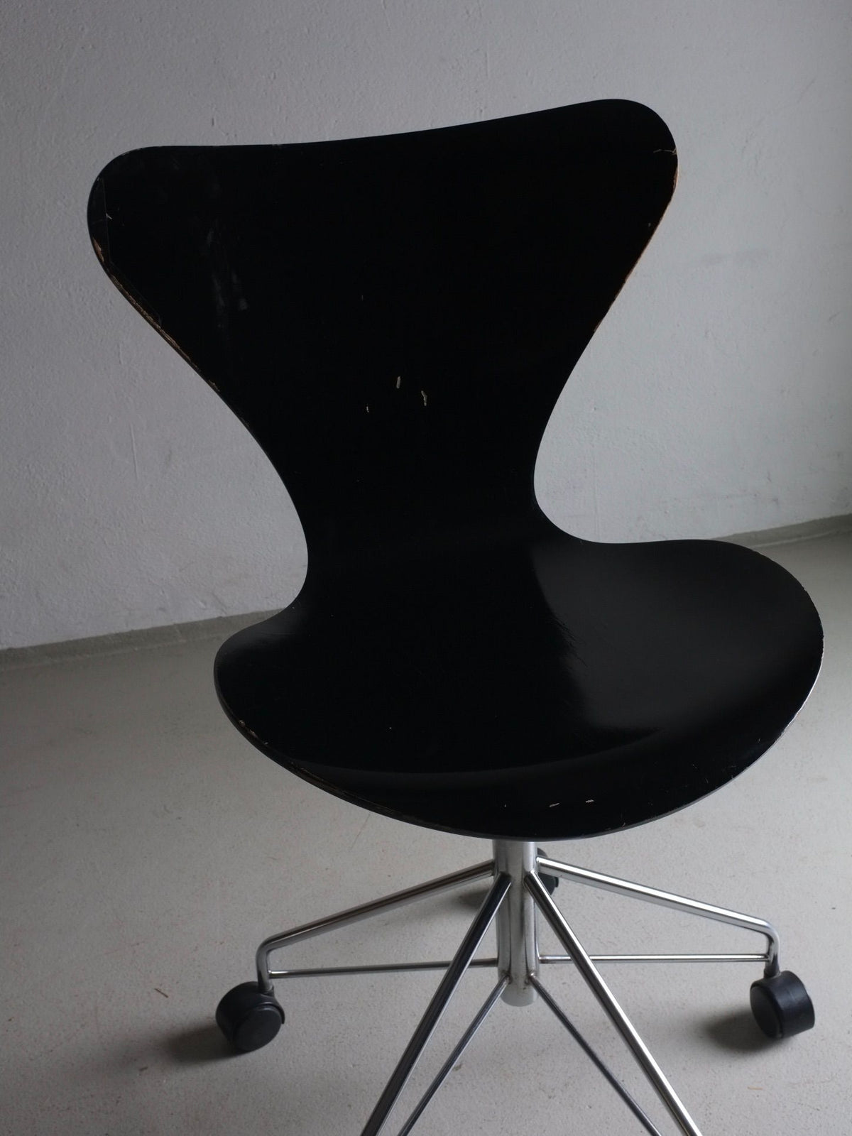 A 3117 Black Swivel Chair by Arne Jacobsen for Fritz Hansen inspired by Arne Jacobsen's designs features a sleek, curved backrest and sits on a chrome swivel base with five casters. The minimalist design by Veter Vintage showcases smooth contours against a plain, light-colored wall and floor.