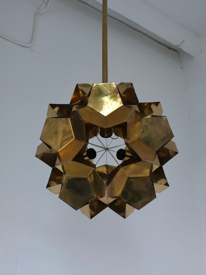 The Brutalist Brass Chandelier by Veter Vintage features a geometric dodecahedron design in solid brass, inspired by 1970s Latvia. This sculptural ceiling pendant is E27 bulb compatible and stands out against a white wall.