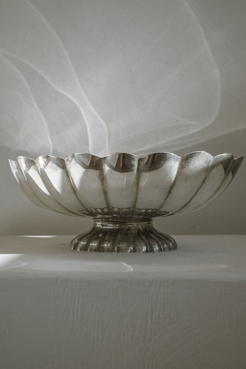 A Silverplated Centerpiece by septembre studios with scalloped edges and a short pedestal base rests on a white textured surface, softly lit to reveal gentle shadows and reflections against a plain background.