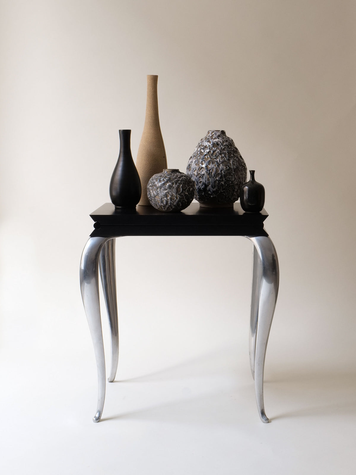 On a modern black table with curved silver legs, five collectable vases of varying shapes in black, gray, and tan sit. Their textured surfaces echo a sleek Louis XV design against a plain cream background. Nearby is the "Lola Mundo Chair" by Philippe Starck (1984) from Collection apart.