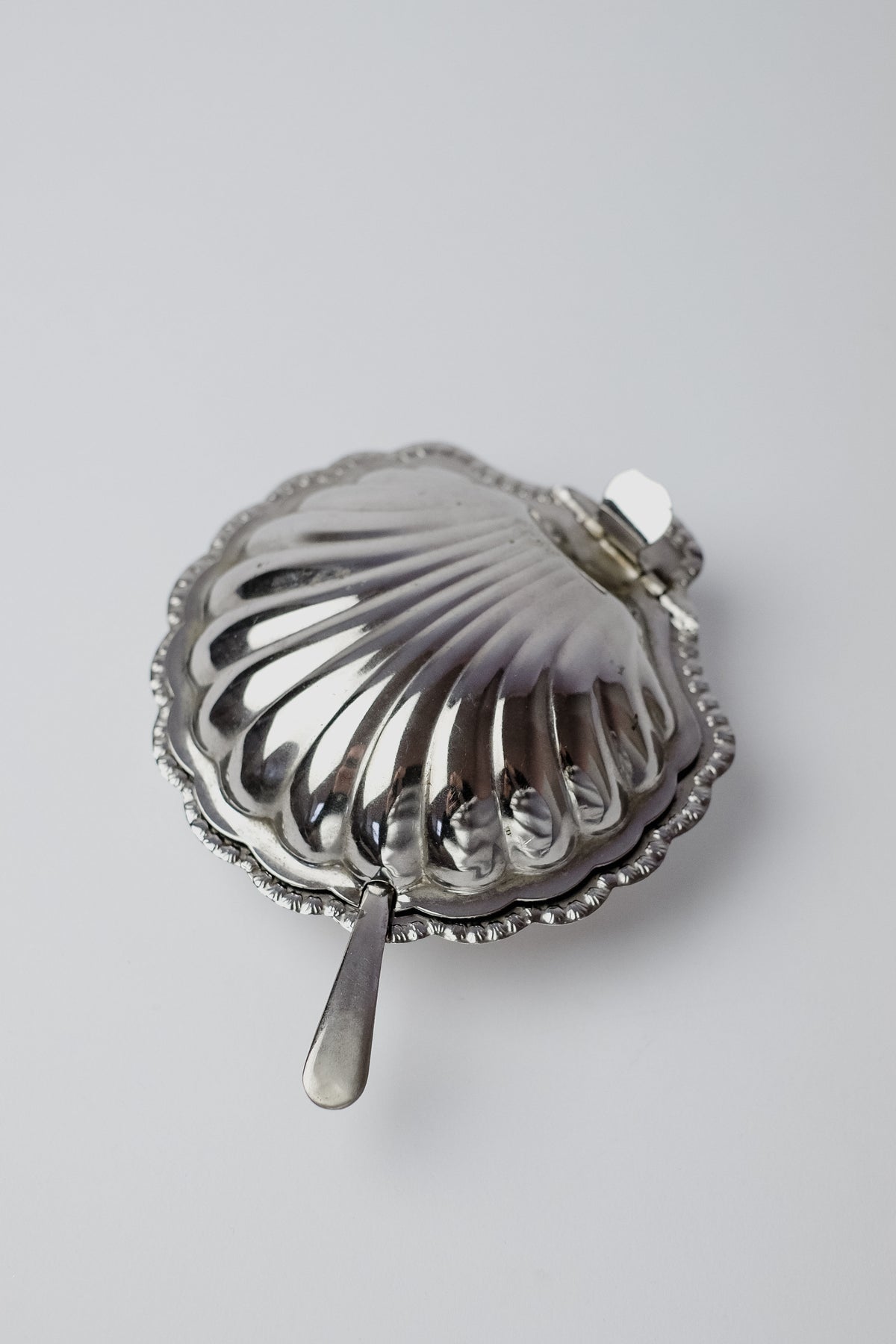 The Shell Shaped Butter Dish with Spoon by septembre studios sits elegantly on a plain white surface. This silver piece, featuring a ribbed texture, scalloped edge, hinged lid, and small spoon, adds understated charm to any dining setting.