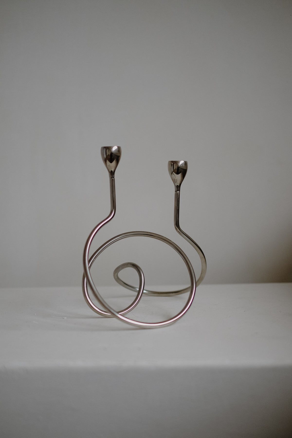 A "Loop" Candle Holder by septembre studios with a minimalist chrome design rests on a white surface against a plain background. It features two spiral arms ending in small candle cups, perfect for dinner candles.