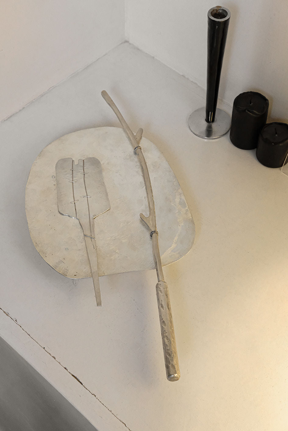 A sleek, minimalist cake server set with a silver-plated base and handle sits on a white surface. Adjacent are three black candles of varying sizes and a tall, black cylindrical candle holder. The setup is modern and understated, reminiscent of a chic Tbilisi, Georgia-inspired aesthetic from Rooms Studio.