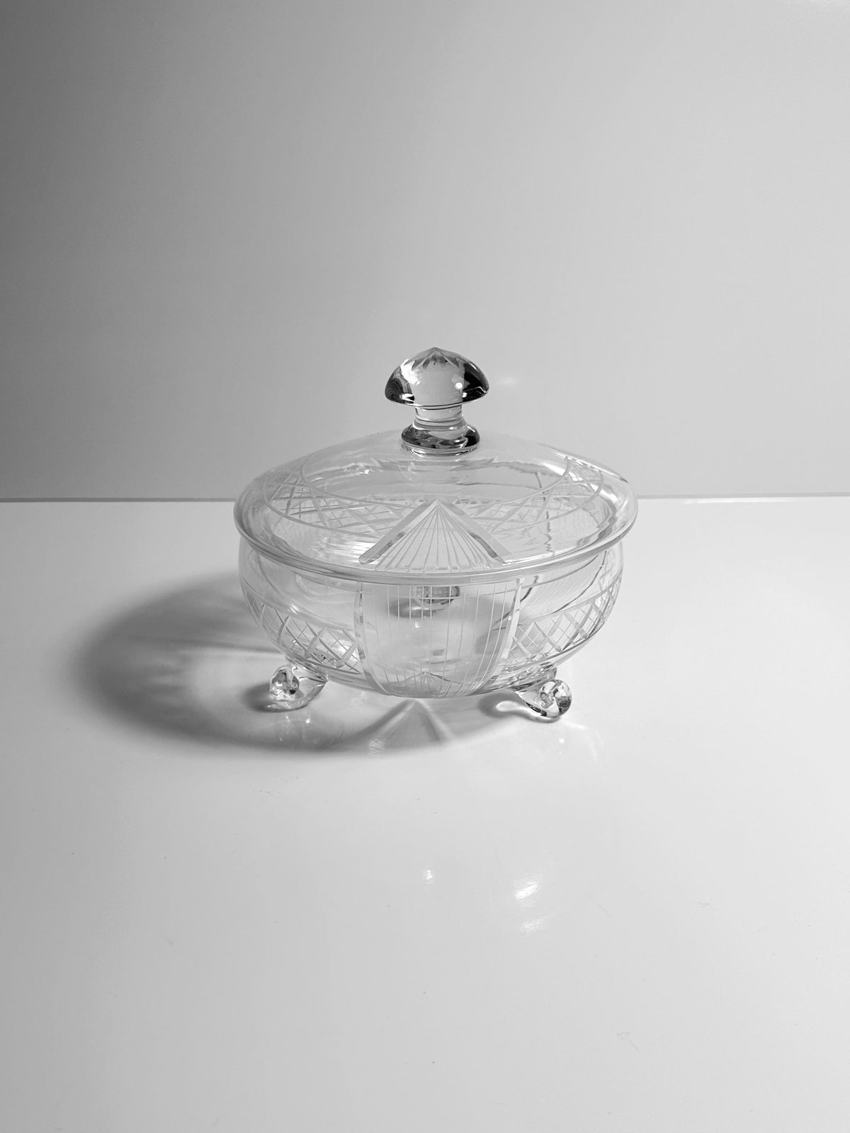 The Art Deco Glass Bonbonnière - 1930 from Dodo Vintage, a clear and intricately designed crystal candy dish, rests on a white surface. It features a decorative pattern and stands on three small feet, casting a subtle shadow.