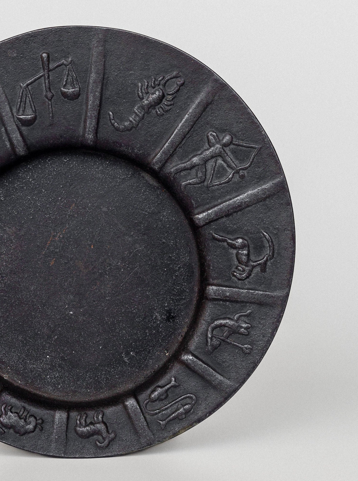 The Art Deco Zodiac Signs Decorative Dish by Spigel is a dark circular plate with vintage iron dish features, showcasing relief carvings of astrological symbols along its rim. The visible symbols include Libra (scale), Scorpio (scorpion), Sagittarius (archer), and Capricorn (sea-goat). This piece embodies Scandinavian vintage design with a plain center.