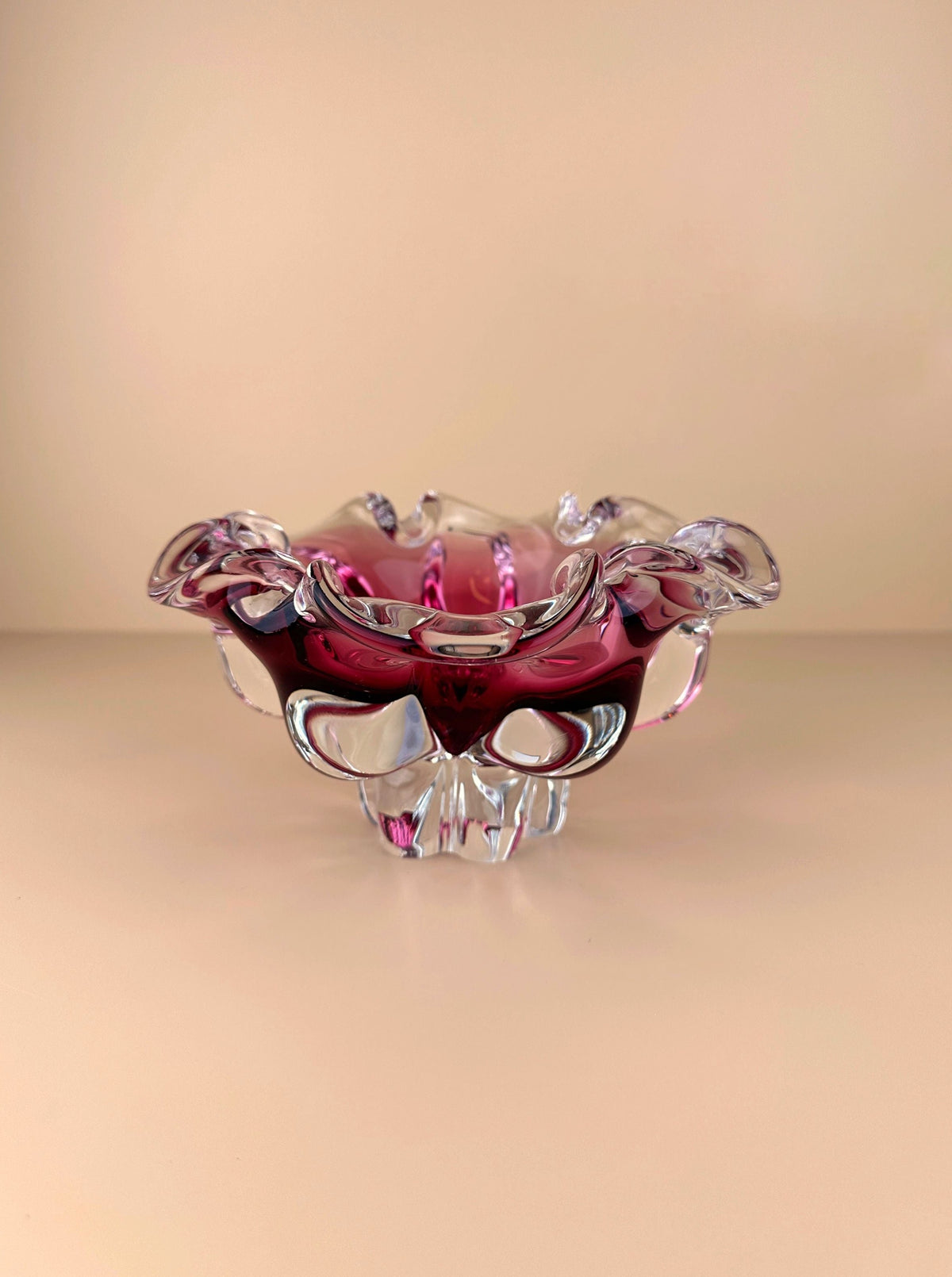 A delicate, translucent pink and clear glass Bohemian Cranberry Red bowl crafted by Josef Hospodka at the Chribska glassworks, with an ornate, ruffled edge design, placed against a soft beige background.