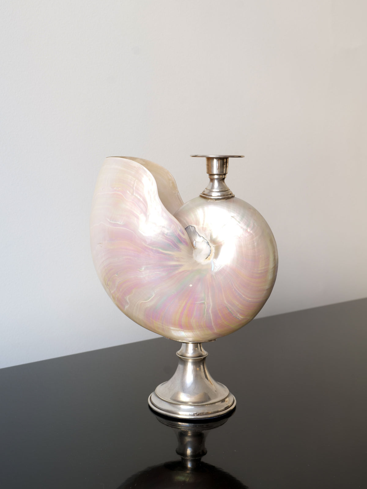 The Nautilus Silver Candle Holder by Collection apart, featuring an iridescent sheen and mounted on a decorative Italian silver base, is elegantly displayed on a reflective black surface against a light gray background.