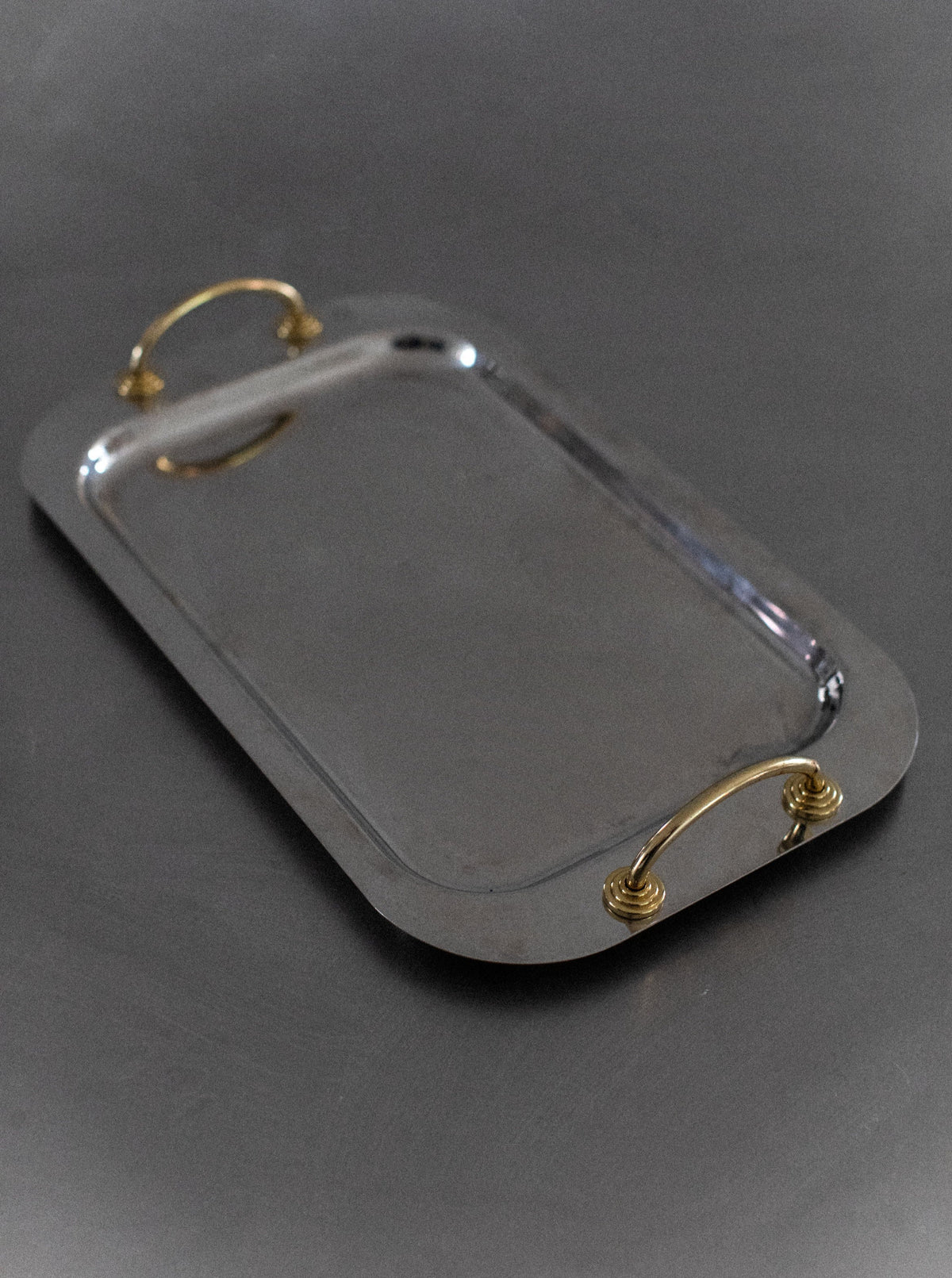 A shiny, rectangular *Out For Lunch* Stainless Steel Tray with rounded corners and two brass handles on the shorter sides is placed on a smooth, dark surface. The tray's reflective finish exudes modern elegance, creating a sleek and contemporary style.