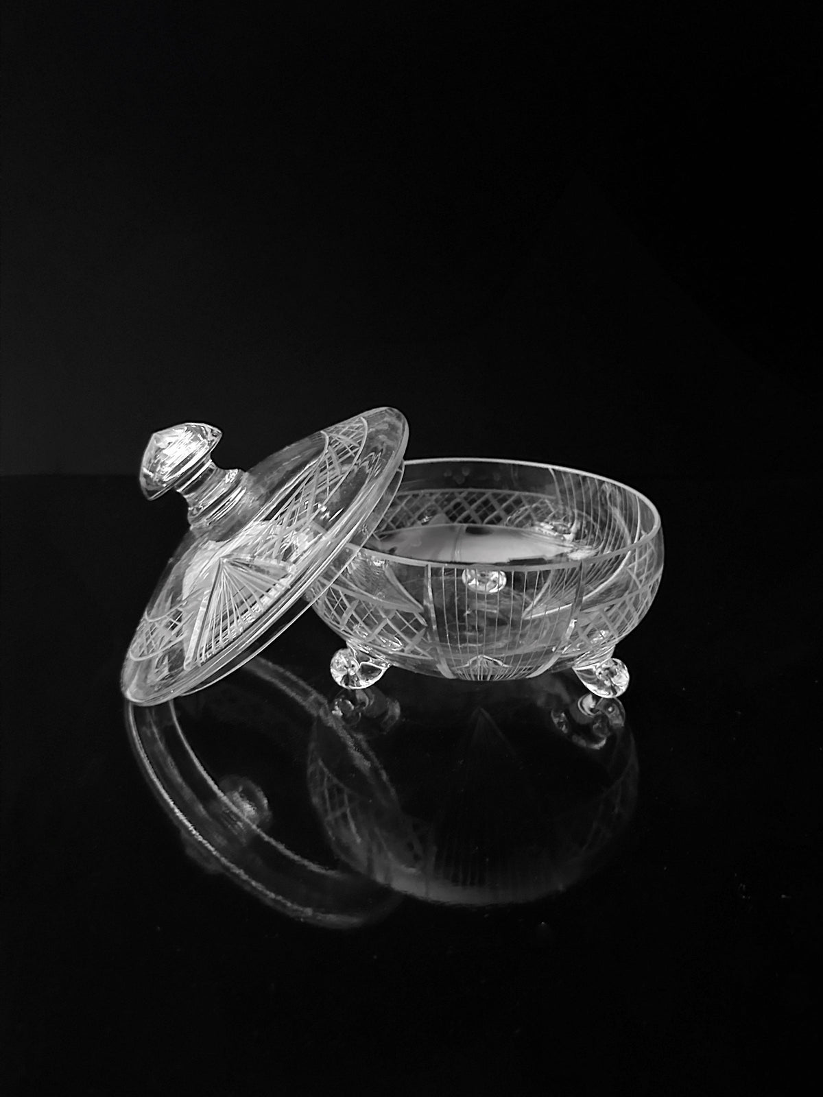 The Art Deco Glass Bonbonnière - 1930 by Dodo Vintage, a handmade piece with intricate patterns, sits open with its round lid resting beside it. The clear crystal reflects light on a glossy black surface, highlighting its ornate Art Deco design and craftsmanship.