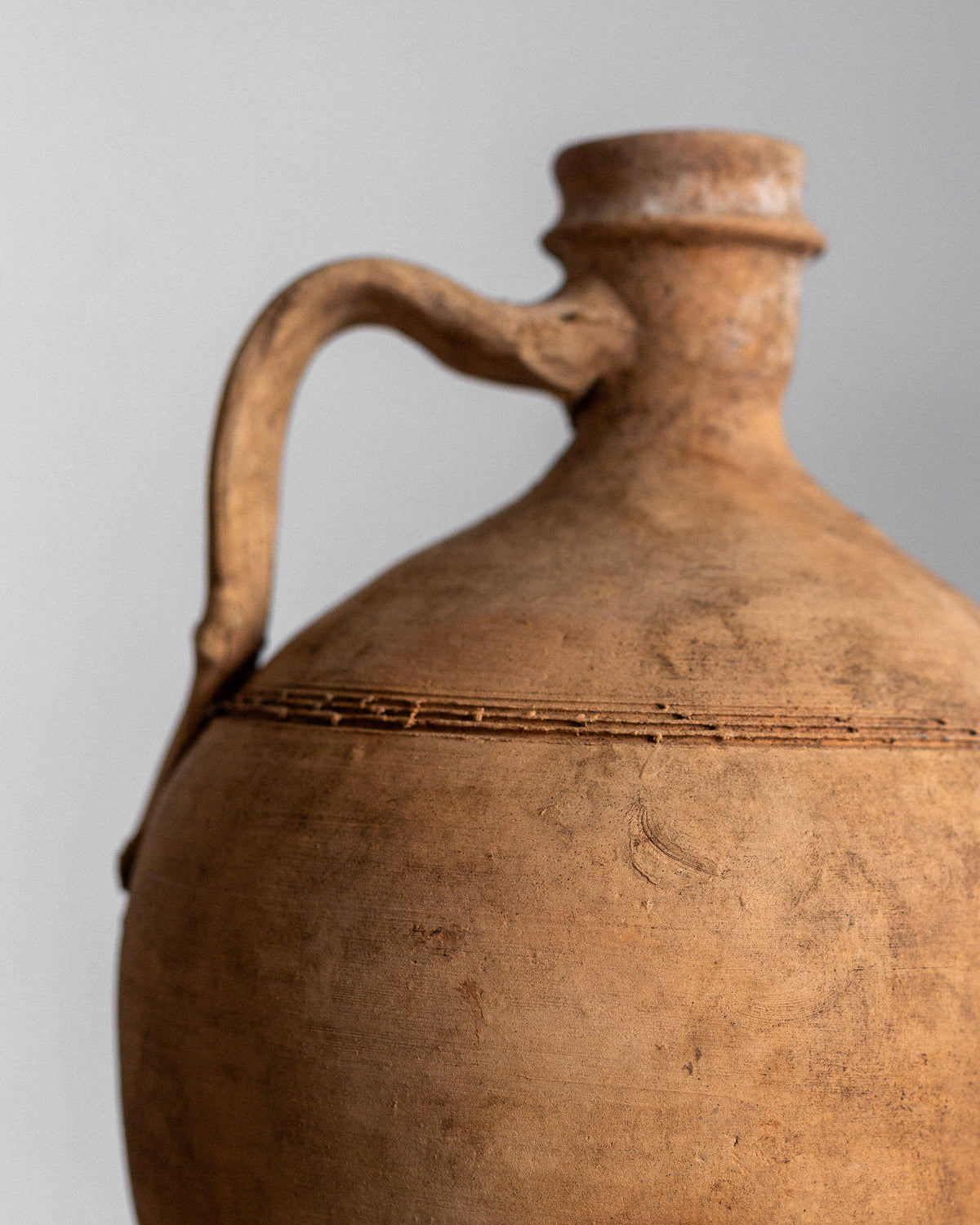A close-up showcases the Spigel Early 20th Century European Olive Jar, featuring a rustic brown finish with a curved handle and narrow neck. The textured surface has horizontal grooves near the top, set against a softly lit background to highlight its earthy tones.