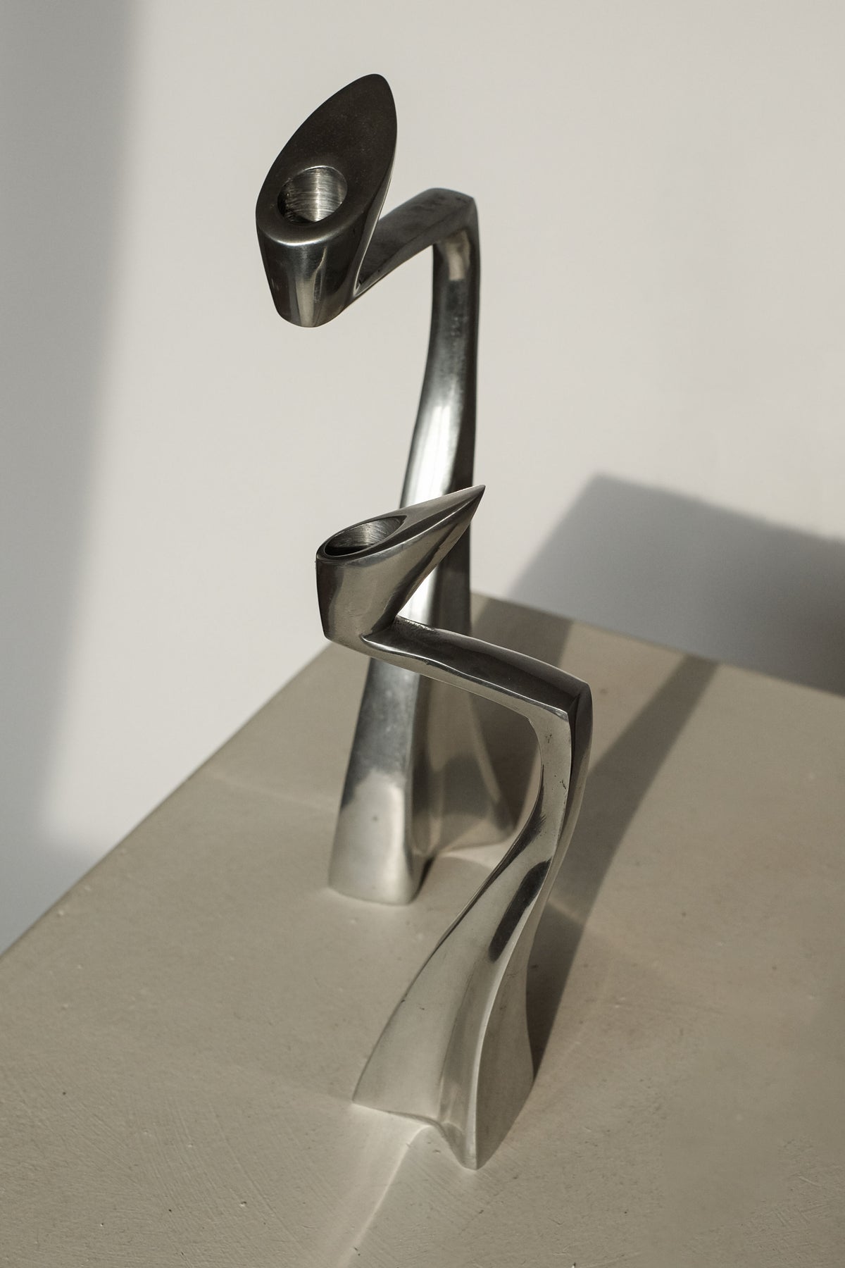 Abstract metal sculpture with intertwined, curved silver forms on a beige surface. Perfect for design enthusiasts, it features smooth reflective surfaces and dynamic twists, casting soft shadows reminiscent of septembre studios' "Pair of Candleholders by Matthew Hilton 80's.