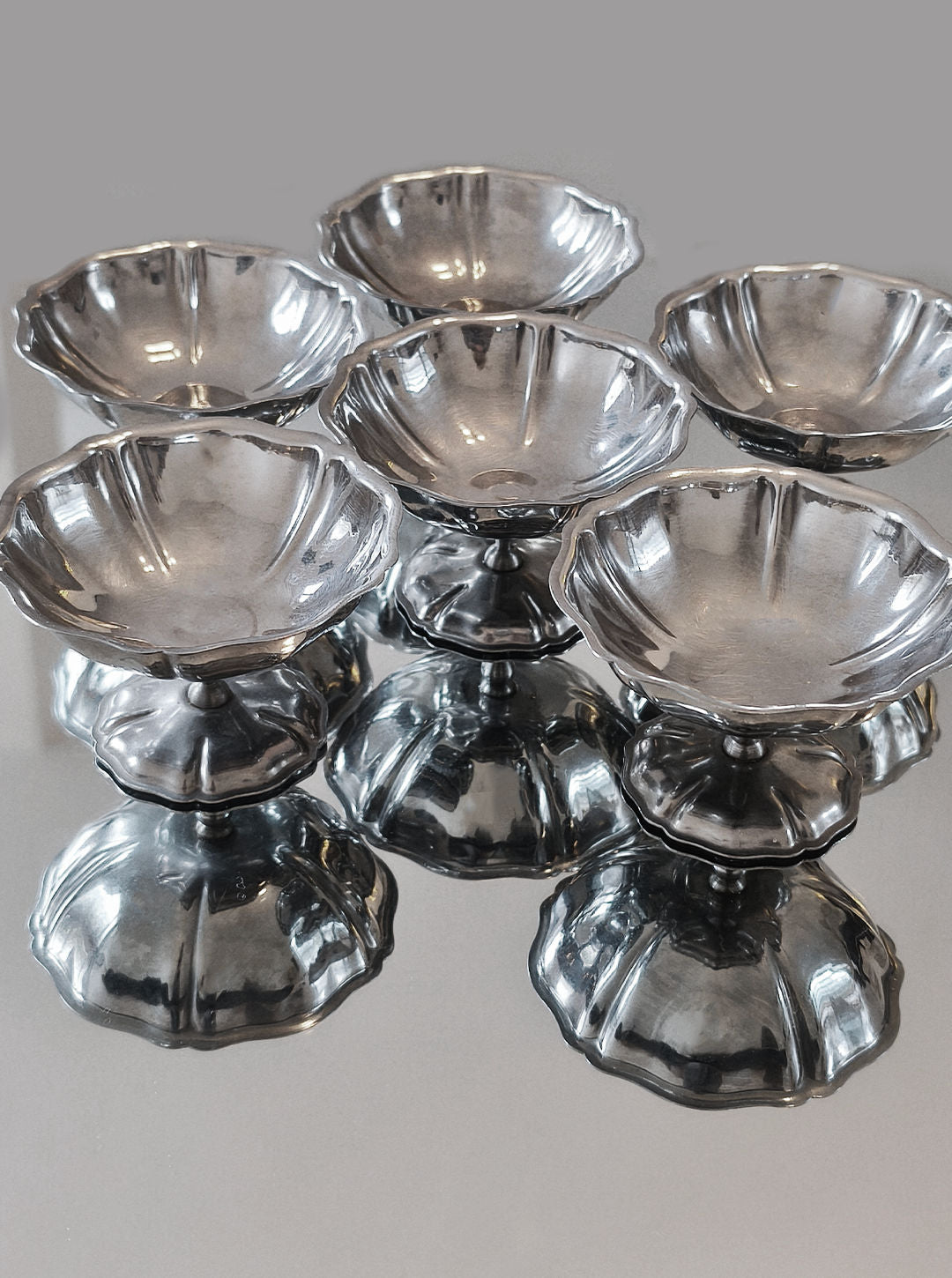 Six polished, stainless steel dessert cups from Les Objoies, each with fluted edges and ornate pedestal bases, are arranged to create a mirrored effect on the reflective surface below.