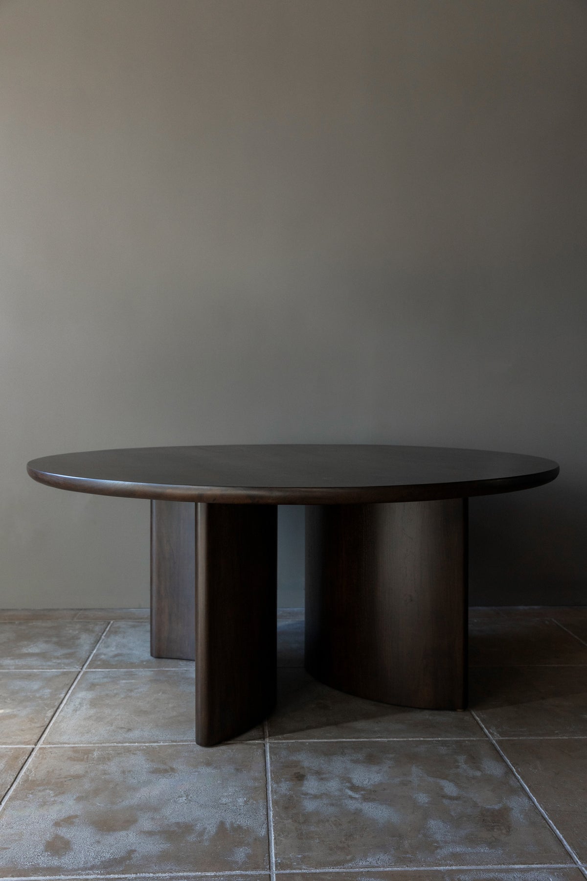 The 'Wright' Dining Table by Studio OSKLO features a round, stained walnut top, three wide curved legs, and stands on large square stone tiles against a plain taupe wall.