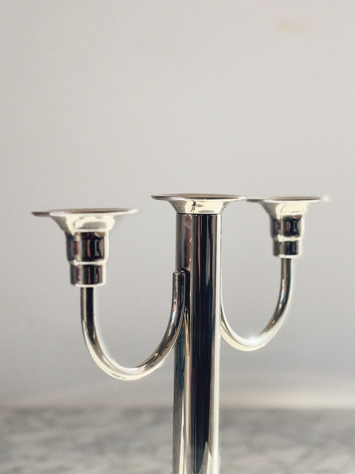 A close-up of the Huerto 3 Arm Metal Candleholder showcases its elegant silver finish and two empty holders, set against a light, blurred background.