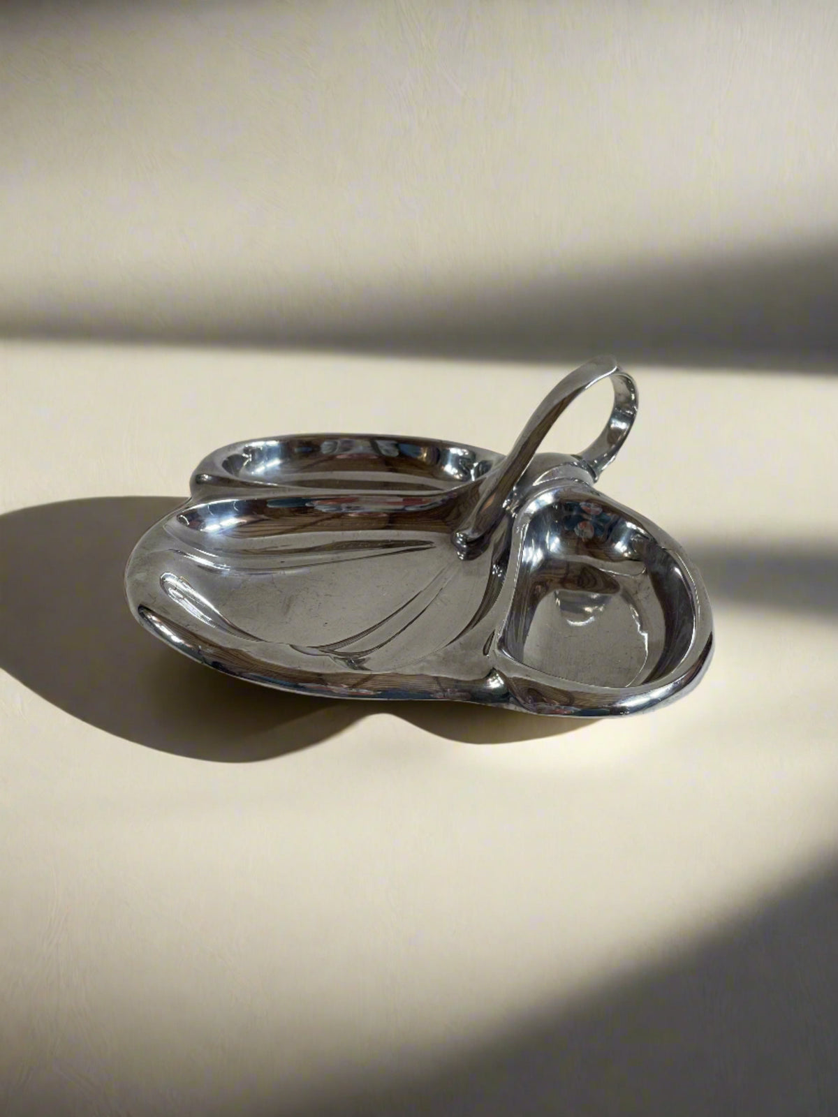 A sleek, modernistic silver Maud Vaughan Vintage Mappin and Webb Silver Plate Serving Dish featuring a three-part design. This smart serving dish has a central, slightly elevated handle for easy carrying, and its three separate compartments are rounded and shiny, reflecting light. The background is plain and light-colored.