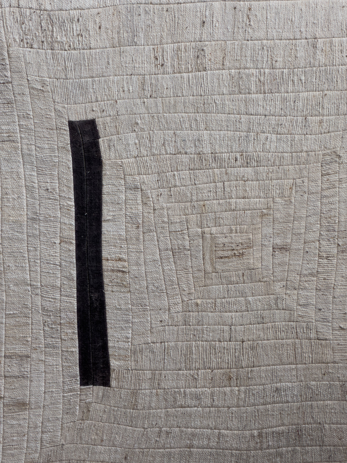 Presenting "to tilt, 2024" by Bec Kirby: This artwork showcases a textured quilt designed with a geometric pattern that evokes the essence of log cabin patchwork. It features concentric rectangles in varying shades of beige and off-white. The design is uniquely disrupted by a single vertical black strip on the left side, creating an unexpected point of interest.