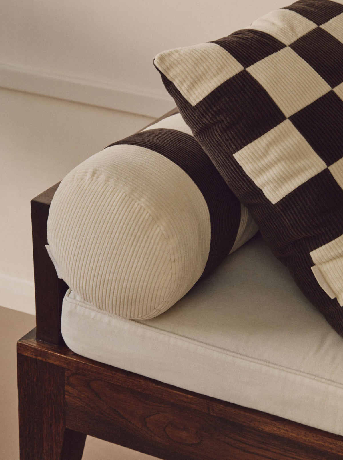 Close-up of a couch with a wooden frame. The armrest features a cylindrical cushion with white and dark brown corduroy stripes, complemented by a black and white checkered throw pillow. Adding character, the sofa includes a vintage-inspired patchwork Handmade UK cushion. The background is softly lit with neutral tones on the wall and floor. Showcased prominently is the AMÉLIORÉ Le Carré - Cotton Corduroy - Chalk/Espresso seat upholstery.
