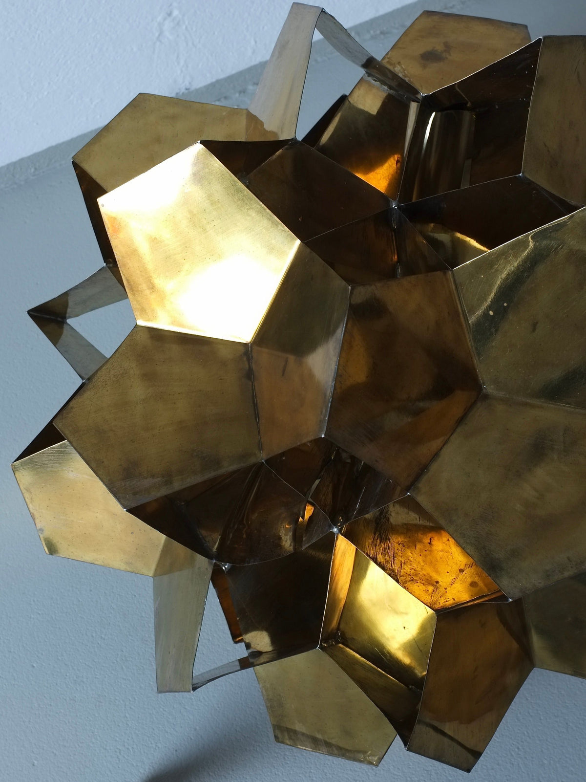 A close-up of the Veter Vintage Brutalist Brass Chandelier, Latvia 70s—interlocking, shiny gold polygons create a sculptural lighting effect against a white and gray backdrop, with light reflecting off its brass surfaces.