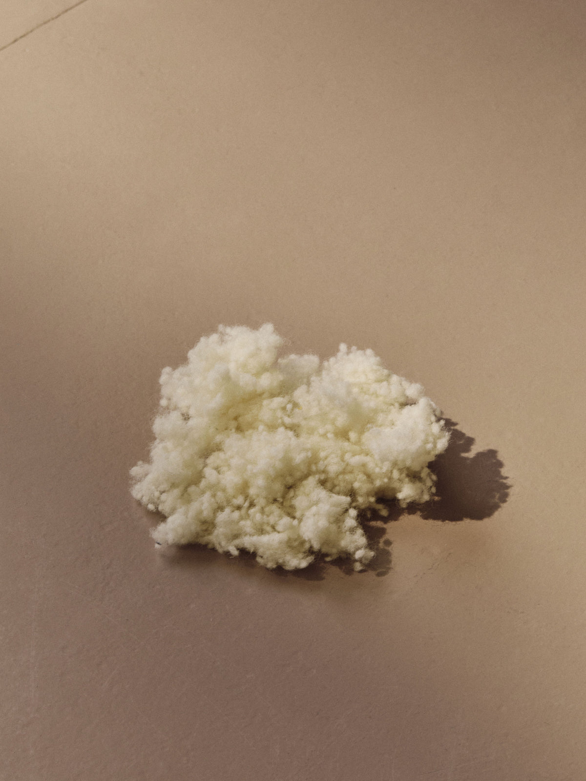 A small, fluffy cloud of cotton-like material, reminiscent of Le Rond - Moyen - Wool Bouclé - Egg White by AMÉLIORÉ, rests on a smooth, beige surface. Loosely clumped together, it casts a gentle shadow to the right due to the lighting.