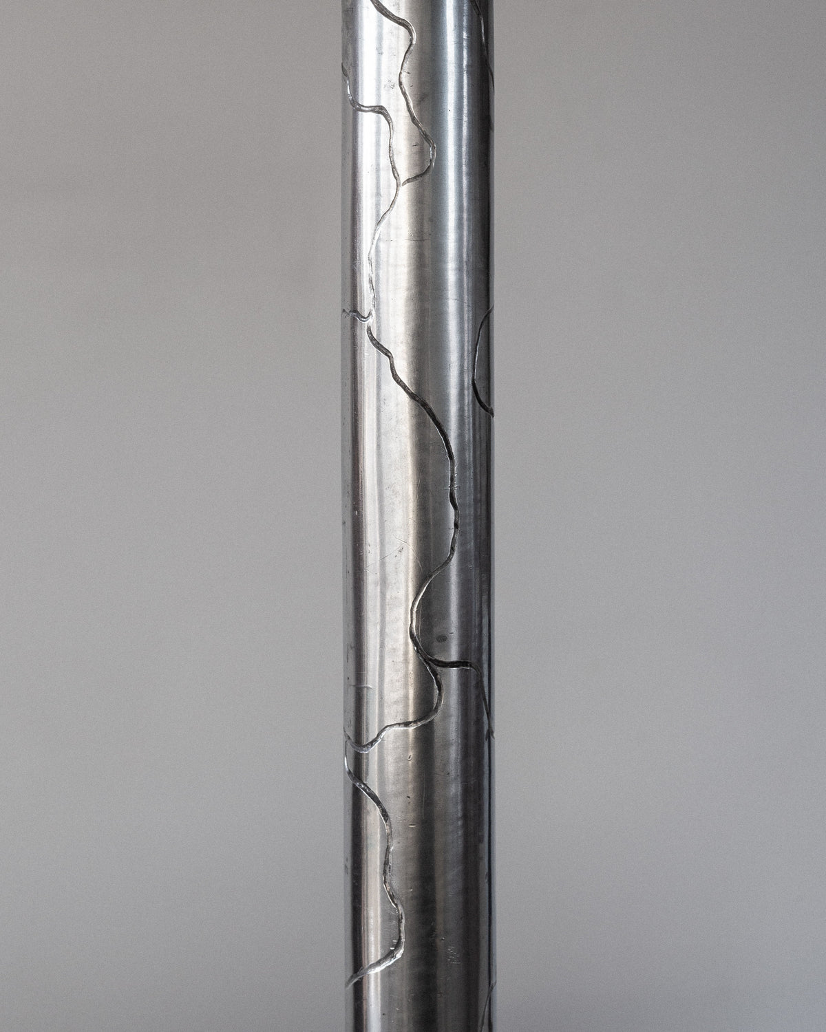 A tall, silver metallic pole features an engraved, irregular vertical line pattern against a gray background, reminiscent of the Art Deco Pewter Lamp (1920s Sweden) by Spigel.