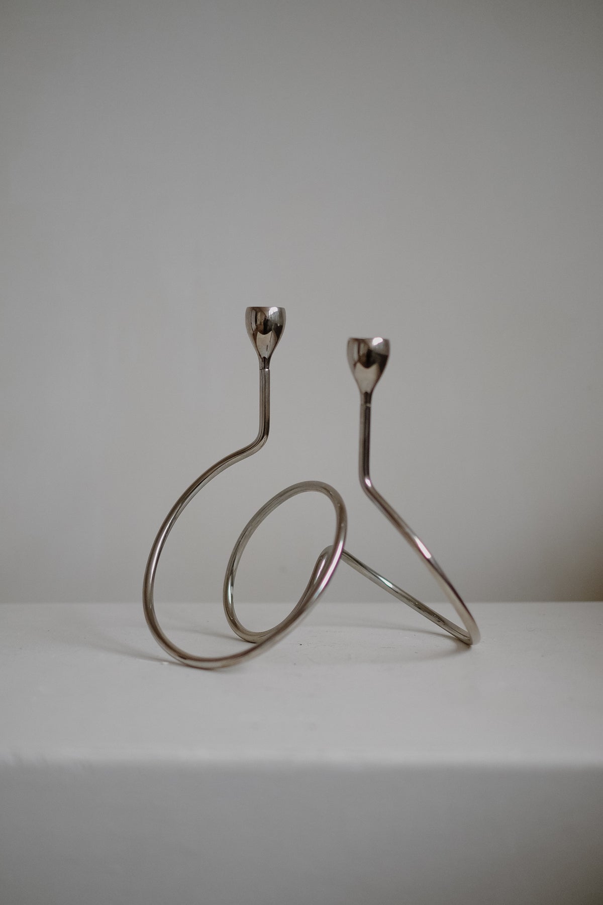The "Loop" Candle Holder by septembre studios features a minimalist chrome design with two holders at varying heights, ideal for dinner candles. It stands elegantly on a white surface with a simple background, showcasing its artistic, abstract shape.