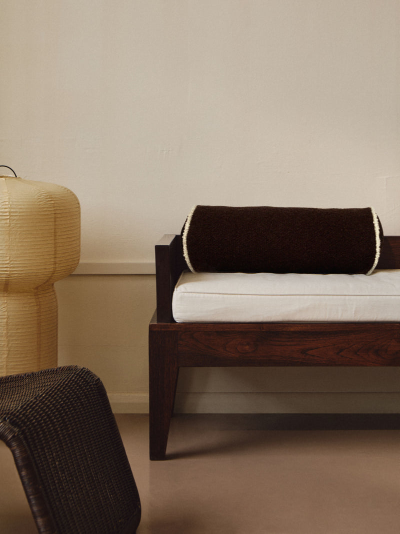 A minimalist room featuring a wooden bench with a white cushion and AMÉLIORÉ's Le Cylindre - Passepoil - Wool Bouclé - Chocolate/Egg White. To the left is a beige, textured lamp, and in the foreground is a wicker chair. The wall and floor are neutral tones, creating a serene atmosphere.