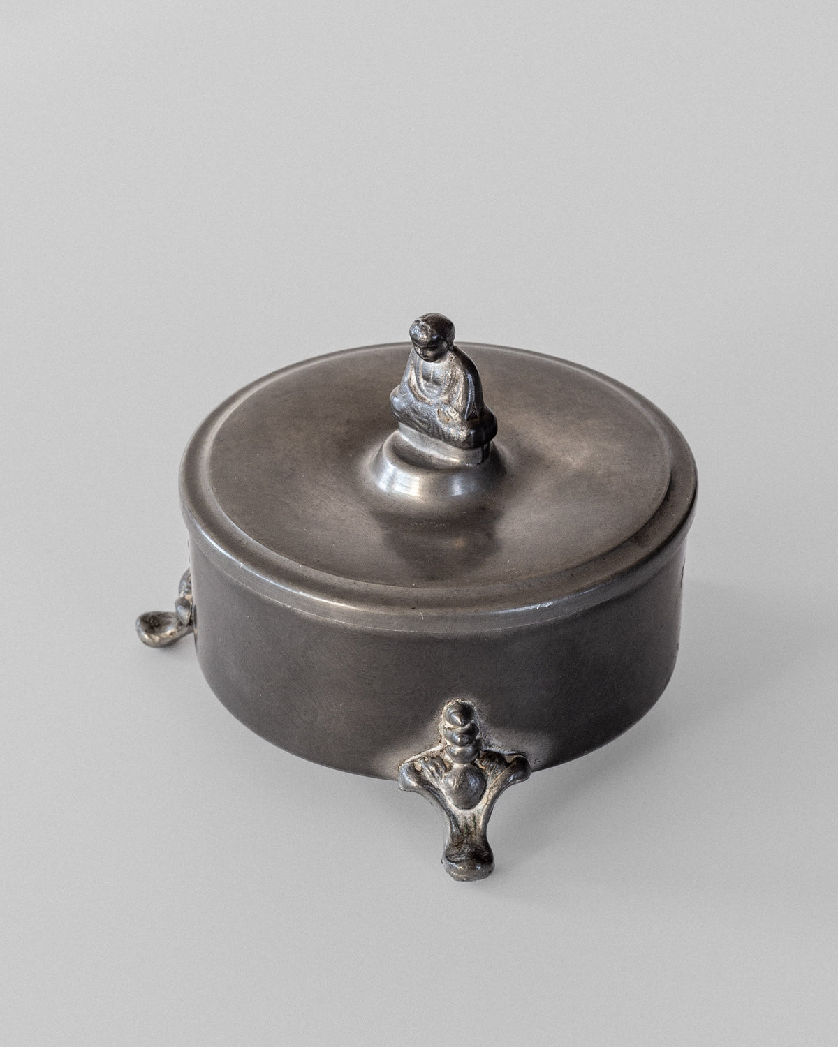The Spigel Art Deco Metal Box from 1934 is a round, intricately designed container with a lid showcasing a small Buddha figure seated atop. Resting on three ornate feet, its sleek metal surface gently reflects light against a simple backdrop.