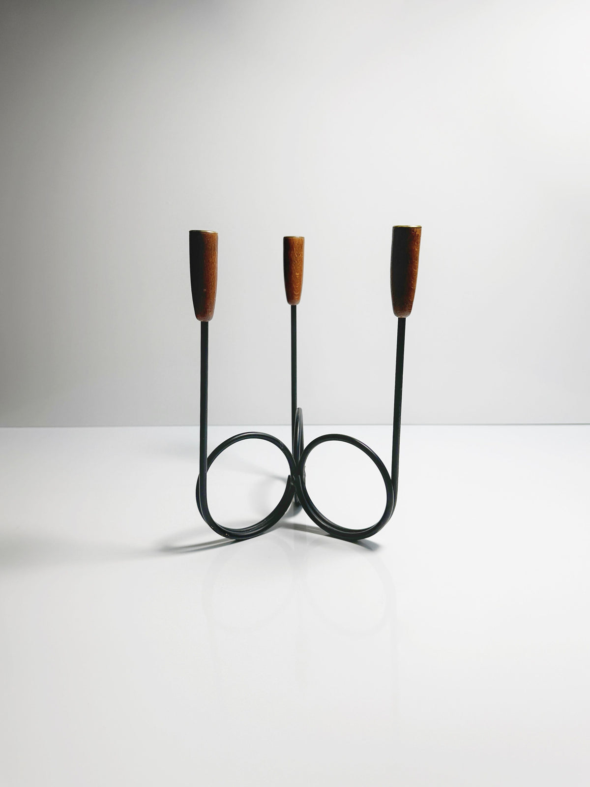 The Danish Candleholder 1960s by Dodo Vintage, a minimalist black wire candelabra with two circular bases and teak ends, designed by Lüthje Denmark, is placed on a reflective white surface with a plain light gray background.