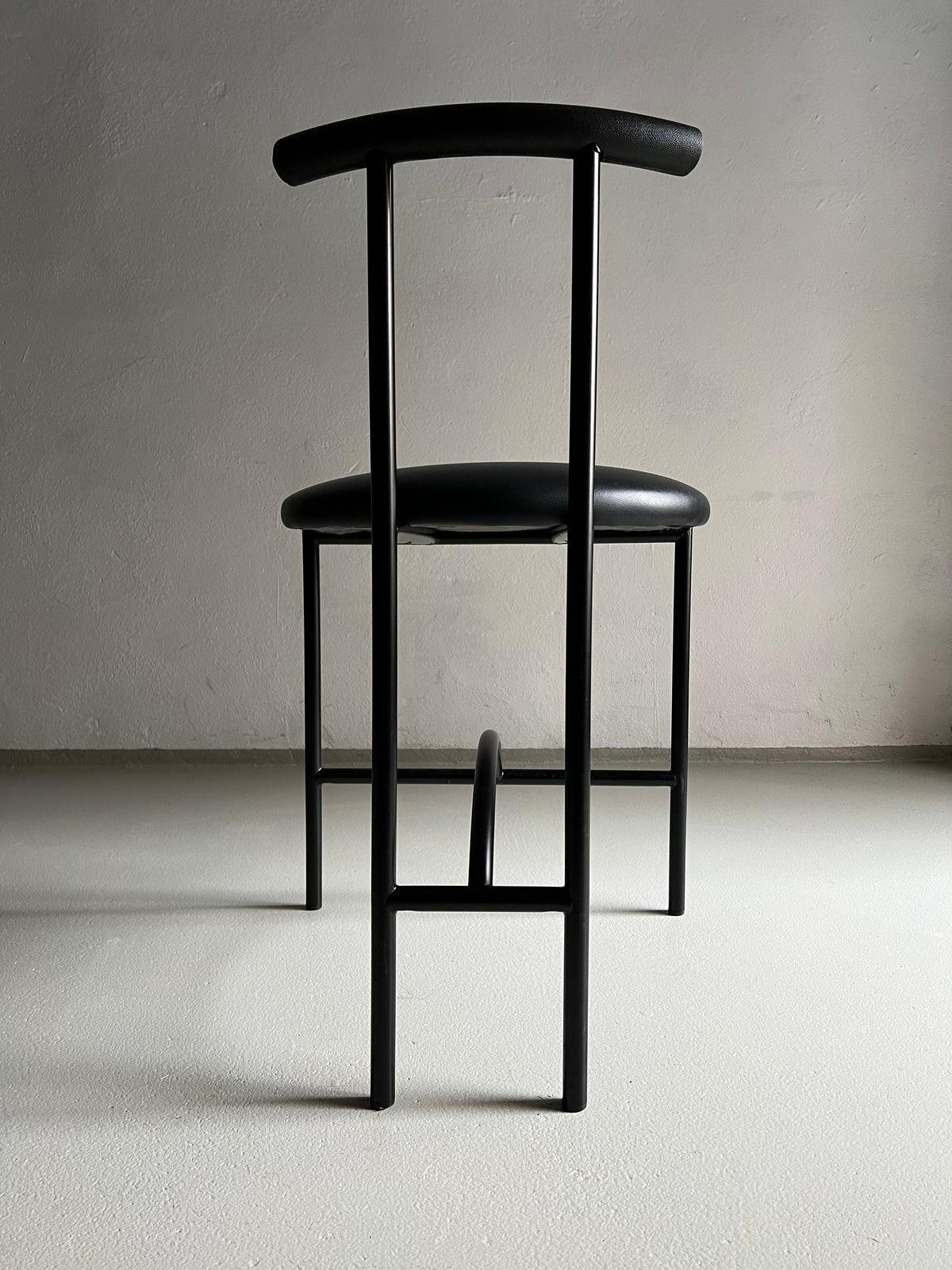 A minimalistic black metal 4 Postmodern "Tokyo" Chairs by Rodney Kinsman | Italy | 1980s with faux leather upholstery is positioned against a plain, light-colored wall. The chair has a simple, modern design with clean lines and an angular frame. Shadows cast subtly on the smooth, light gray floor beneath it.
