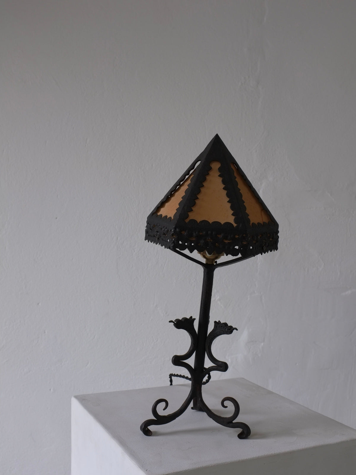 The Veter Vintage Wrought Iron Dragon Table Lamp, Italy 1910s, with dragon accents and a triangular cream silk shade, is displayed on a white pedestal against a plain light gray wall.