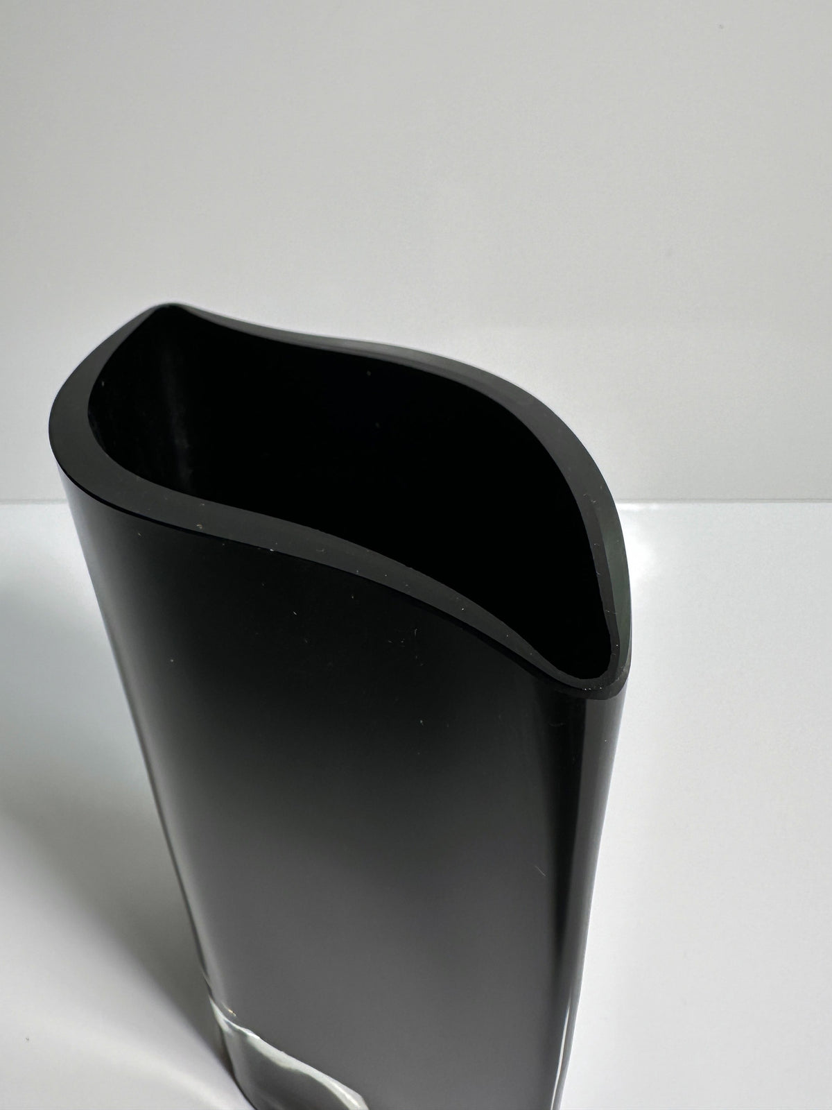 A sleek, modern black glass vase by Kaj Franck from Dodo Vintage stands on a white surface. The Nuutajärvi Notjö vase features a curved, asymmetrical shape with a glossy finish and a wide opening at the top that narrows slightly towards the base. The background is plain and light-colored.