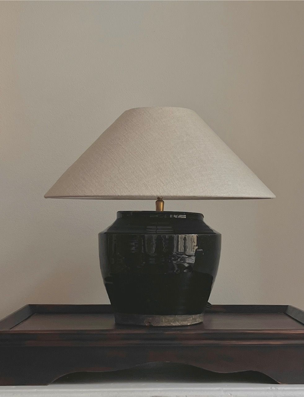 The Antique Glazed Lamp by Médecine features a black ceramic base and a wide beige lampshade. It sits on a dark wooden table, accompanied by a black textile cord, against a plain light-colored wall.