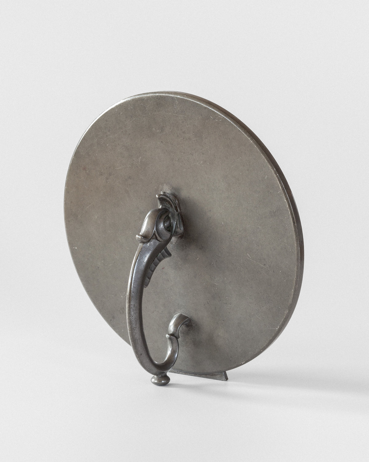 A vintage Spigel Art Deco Metal Mirror in antique condition showcases a round reflective surface and an ornate handle. Set against a plain backdrop, it is slightly tilted to reveal its side profile and intricate design details.