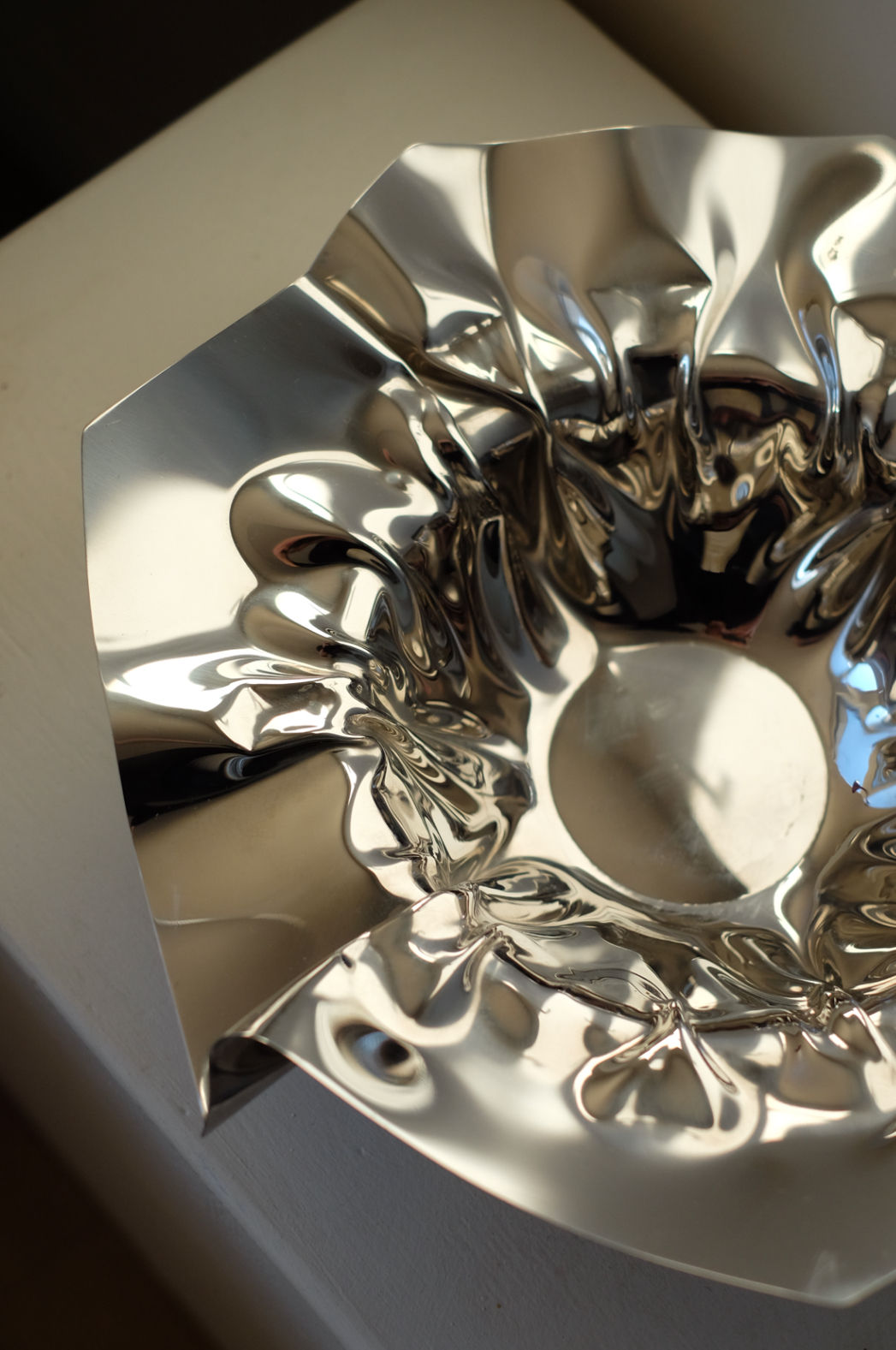 A Folded Chrome Bowl by septembre studios, featuring a shiny, reflective metal finish and a crumpled, irregular surface, sits on a light-colored interior surface. Its textured design casts soft reflections and shadows, creating an abstract and artistic effect.