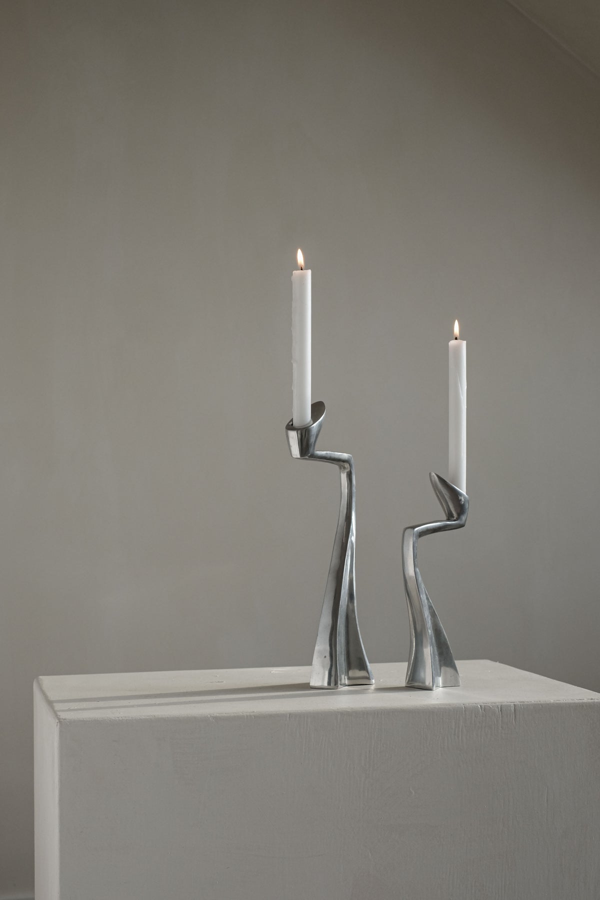 Two lit white candles sit in a Pair of Candleholders by Matthew Hilton 80's from septembre studios, placed on a minimalist white pedestal against a neutral wall, where the sleek design contrasts beautifully with the candlelight for an elegant and calming ambiance.