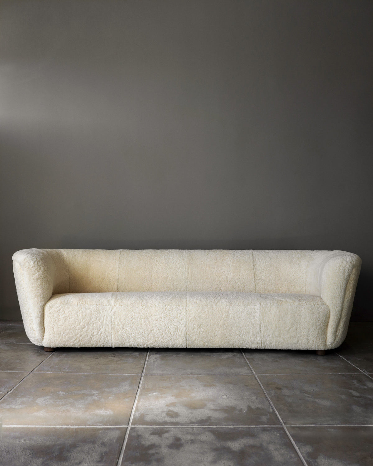 A Fritz Hansen Sofa in Shearling by Studio OSKLO, featuring a cream, textured finish, sits against a plain gray wall on a tiled concrete floor.